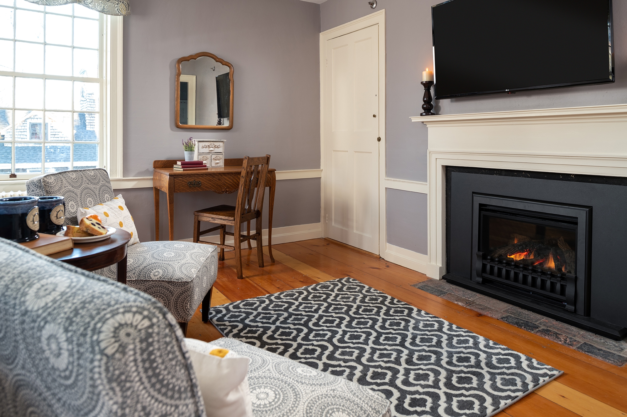 A cozy seating area in a guestroom featuring a black gas fireplace with a white mantel and mounted TV, a small wooden writing desk with a chair, and patterned armchairs on wide-plank wood floors.