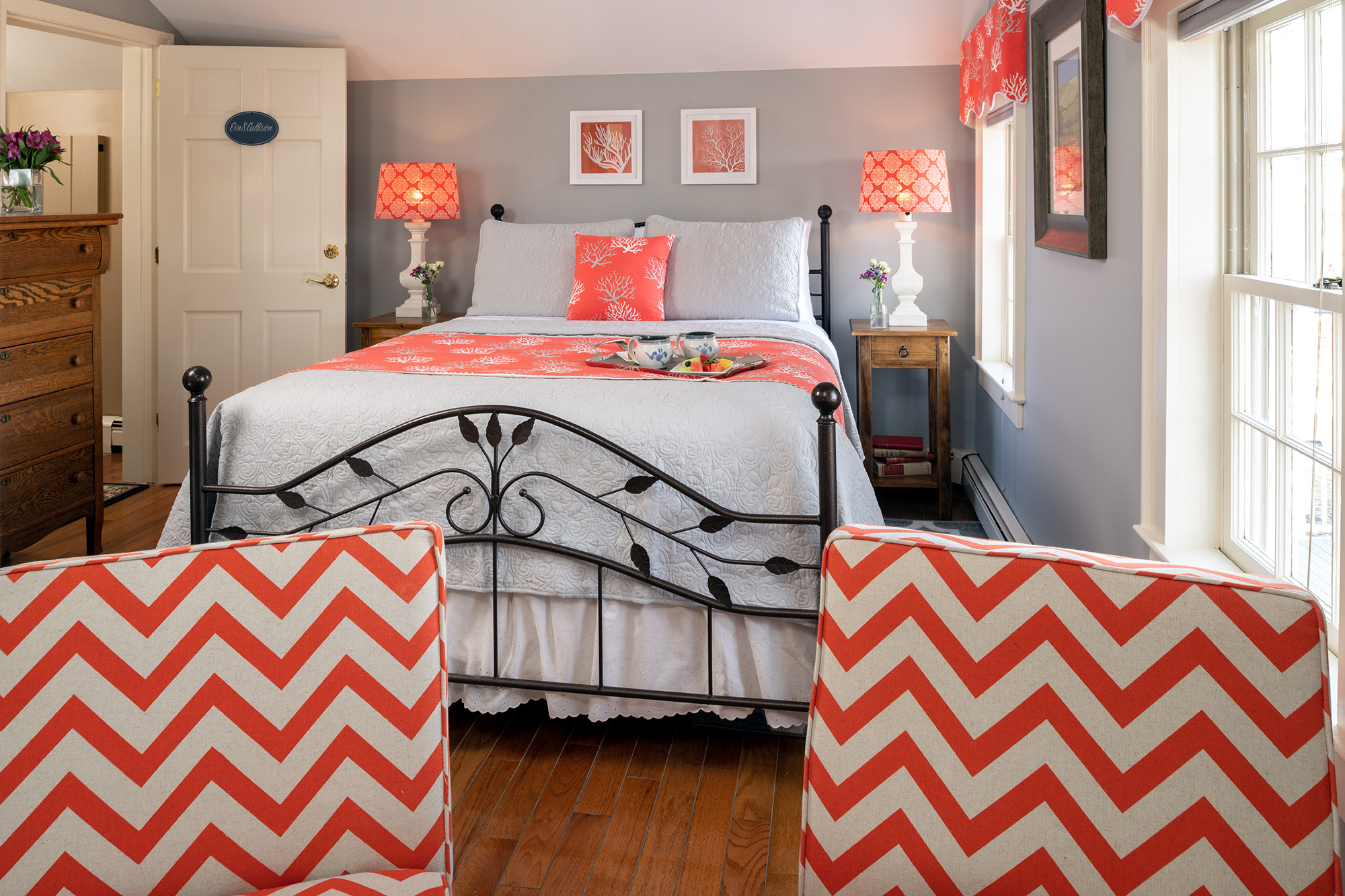 A vibrant guestroom with slate gray walls and hardwood floors, featuring a black wrought-iron bed with coral and white linens, matching coral patterned lamps, and two chevron-patterned chairs in the foreground.