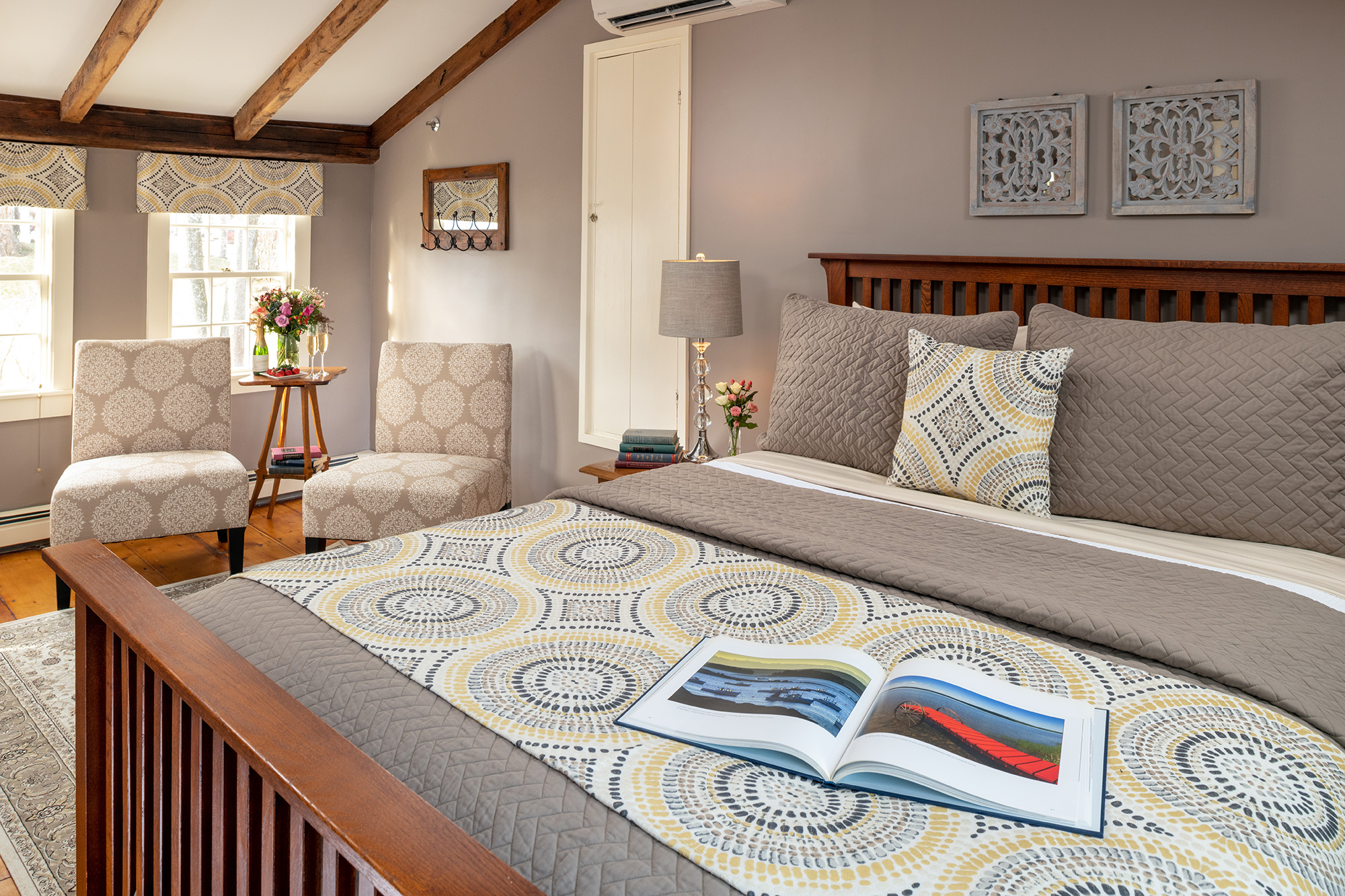 A serene guestroom with taupe walls and a vaulted ceiling with exposed wood beams, featuring a queen-size wooden bed with grey and yellow geometric linens and a small seating area with two patterned slipper chairs by the window.