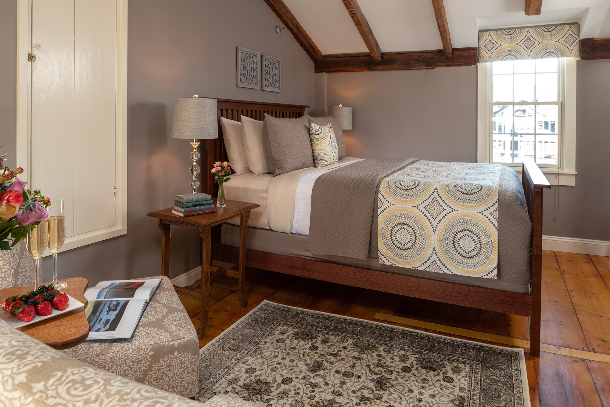 A warm and inviting guestroom featuring a wooden bed with grey and yellow geometric linens, set against taupe walls and wide-plank wood floors. The room is accented by vaulted ceilings with exposed wood beams and a cozy patterned area rug.
