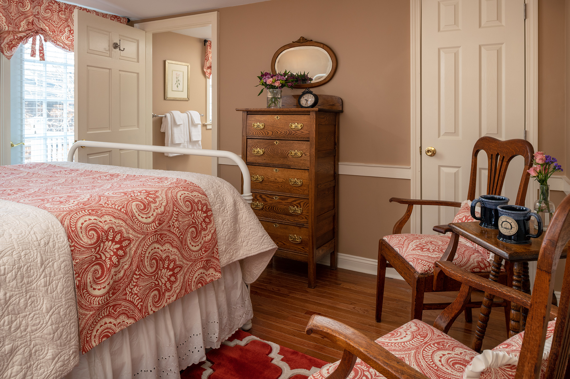 A quaint, traditional guestroom with warm tan walls and hardwood floors, featuring a white iron bed with coral paisley linens, a vintage tallboy dresser, and a seating area with two wooden armchairs upholstered in matching coral fabric.