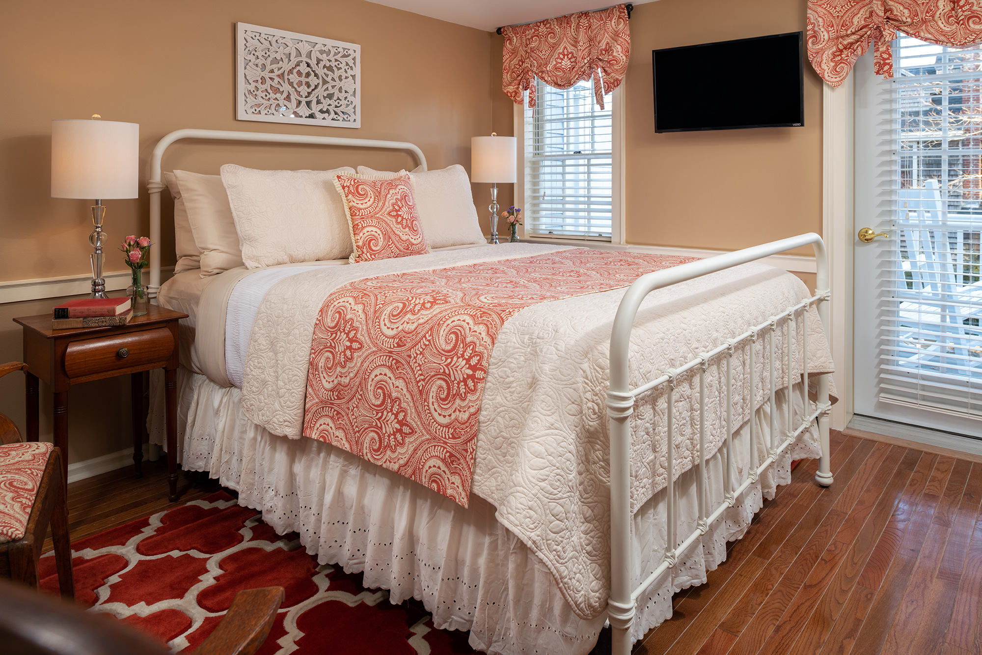 A charming guestroom featuring a white iron bed with coral and cream paisley linens, warm tan walls, and hardwood floors. The room includes a mounted flat-screen TV and a glass door leading to a private outdoor deck.