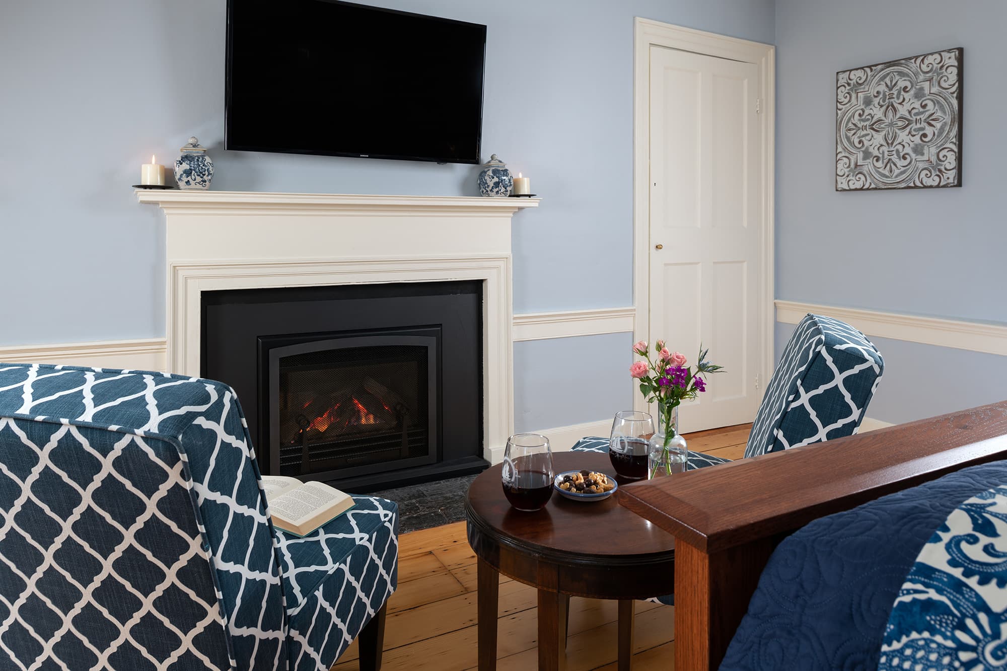 A cozy sitting area in a light blue guestroom featuring a black gas fireplace with a white mantel, a mounted TV, and two teal diamond-patterned chairs. A small wooden table in the foreground holds wine and snacks.