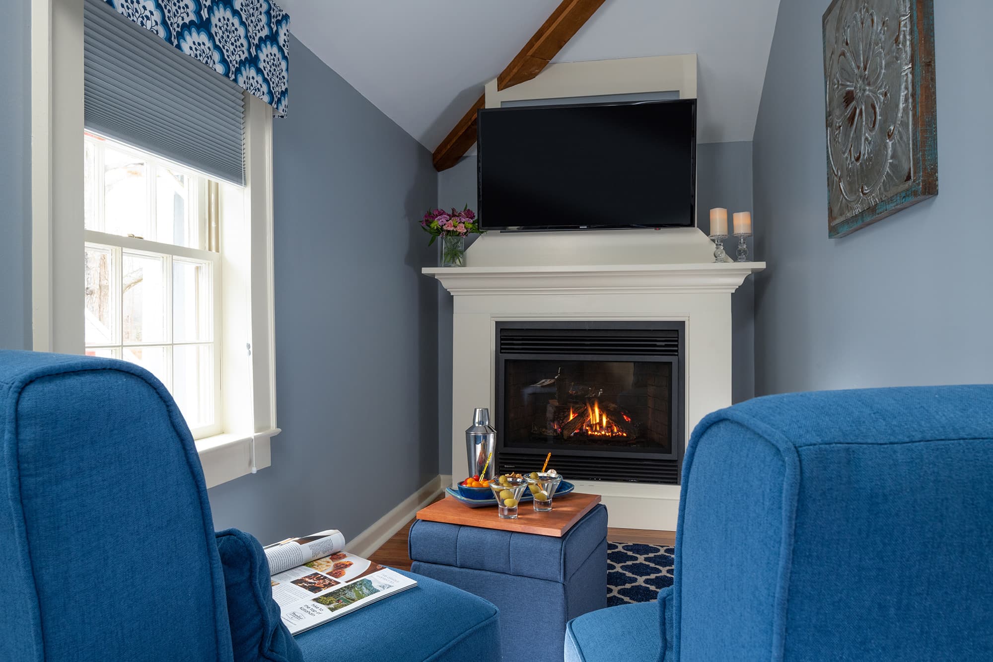 A cozy nook in a light blue guestroom featuring a white gas fireplace with a mounted TV above, two bright blue armchairs, and a matching storage ottoman holding a tray with drinks.