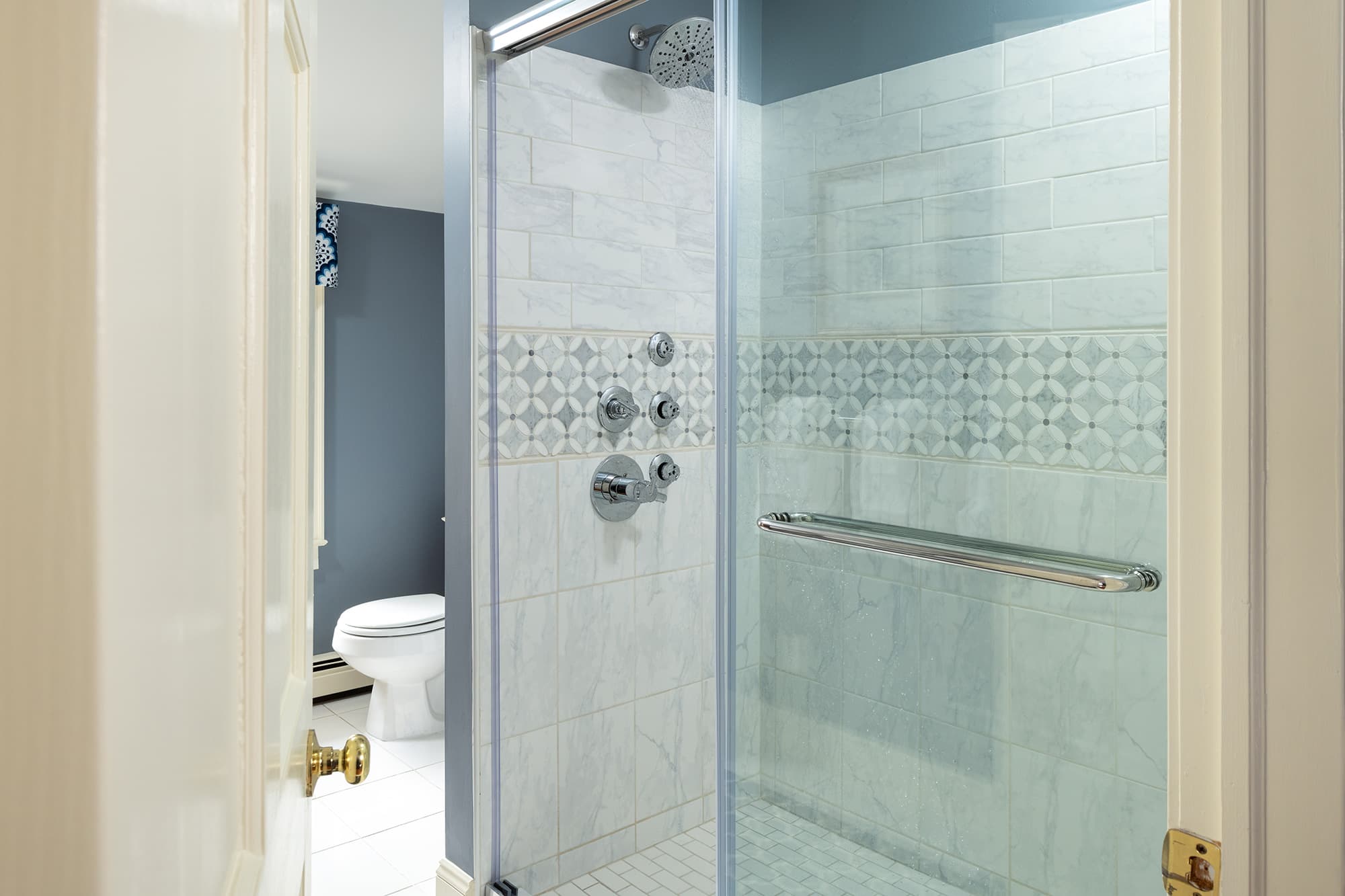 A bright en-suite bathroom featuring a glass-enclosed walk-in shower with white subway tiles, a decorative teal patterned tile border, and luxury shower fixtures against slate blue walls.