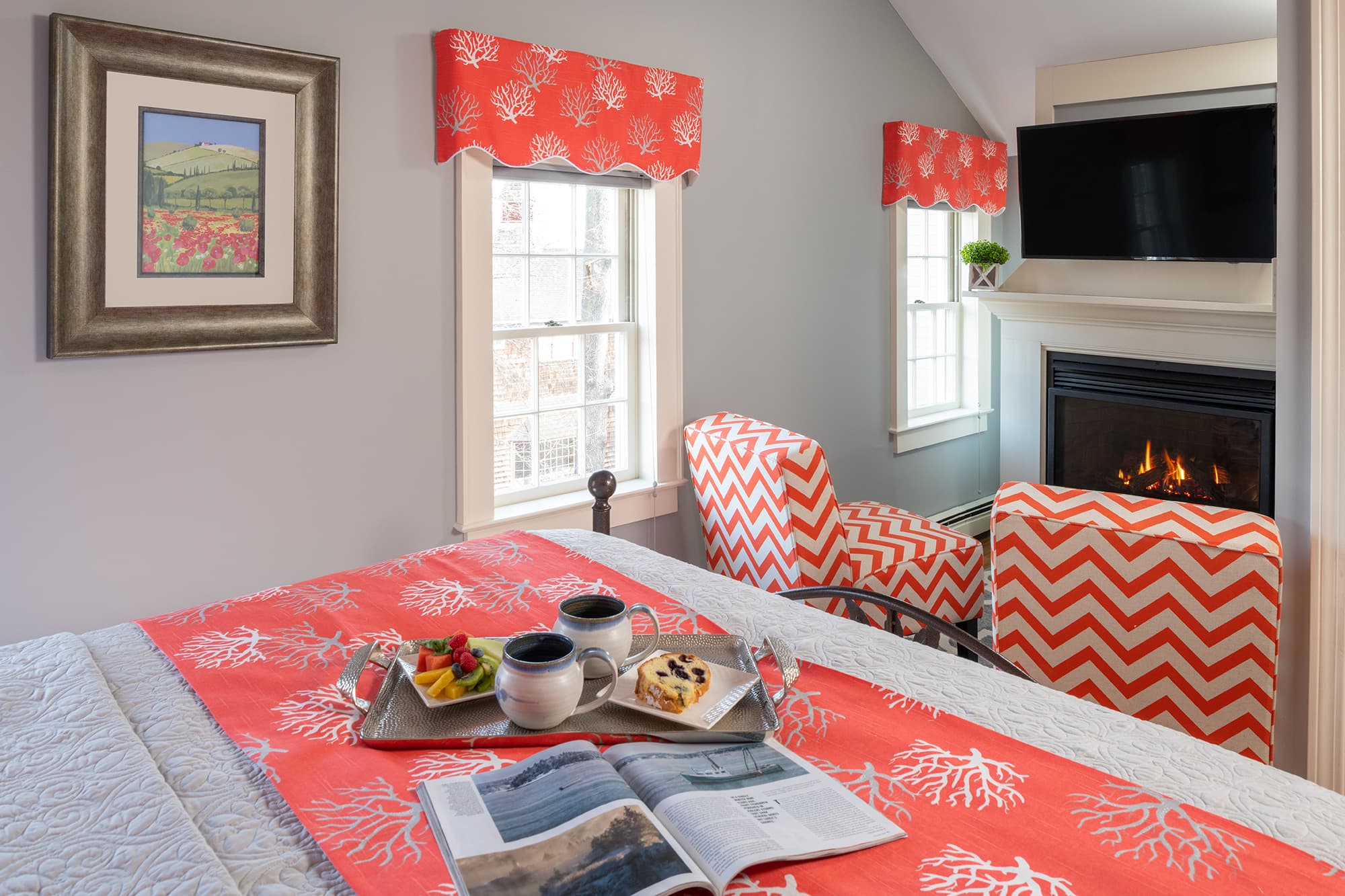 A cozy guestroom seating area featuring two coral-and-white chevron armchairs positioned in front of a warm gas fireplace with a mounted TV. A breakfast tray with coffee and fruit rests on the bed in the foreground.