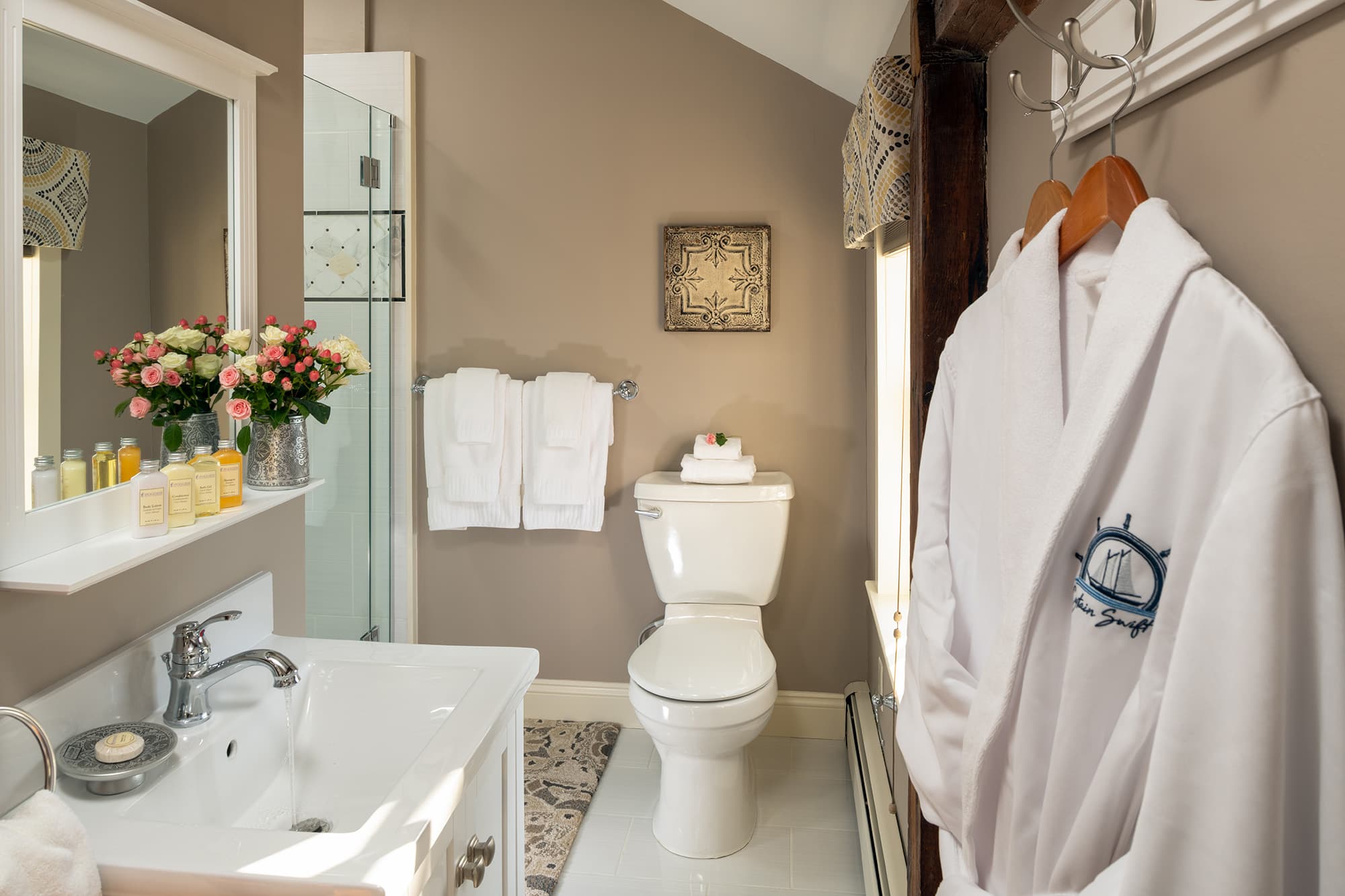 A clean and bright en-suite bathroom with taupe walls, featuring a white vanity and sink, a white plush robe hanging in the foreground, and a glass-enclosed shower in the background.