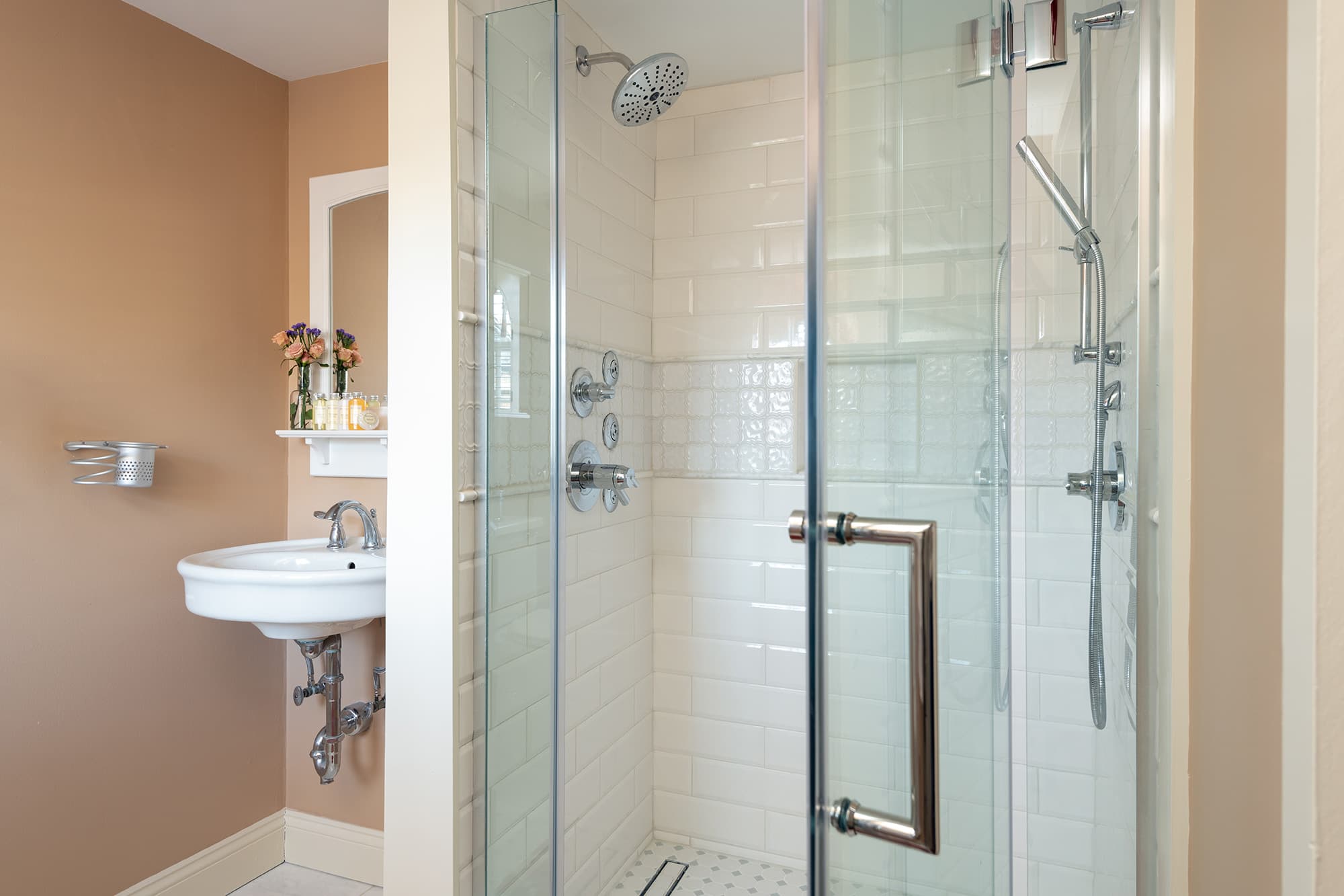 A bright en-suite bathroom featuring a modern glass-enclosed walk-in shower with white subway tiles and multi-jet body sprays, located next to a white pedestal sink against warm tan walls.