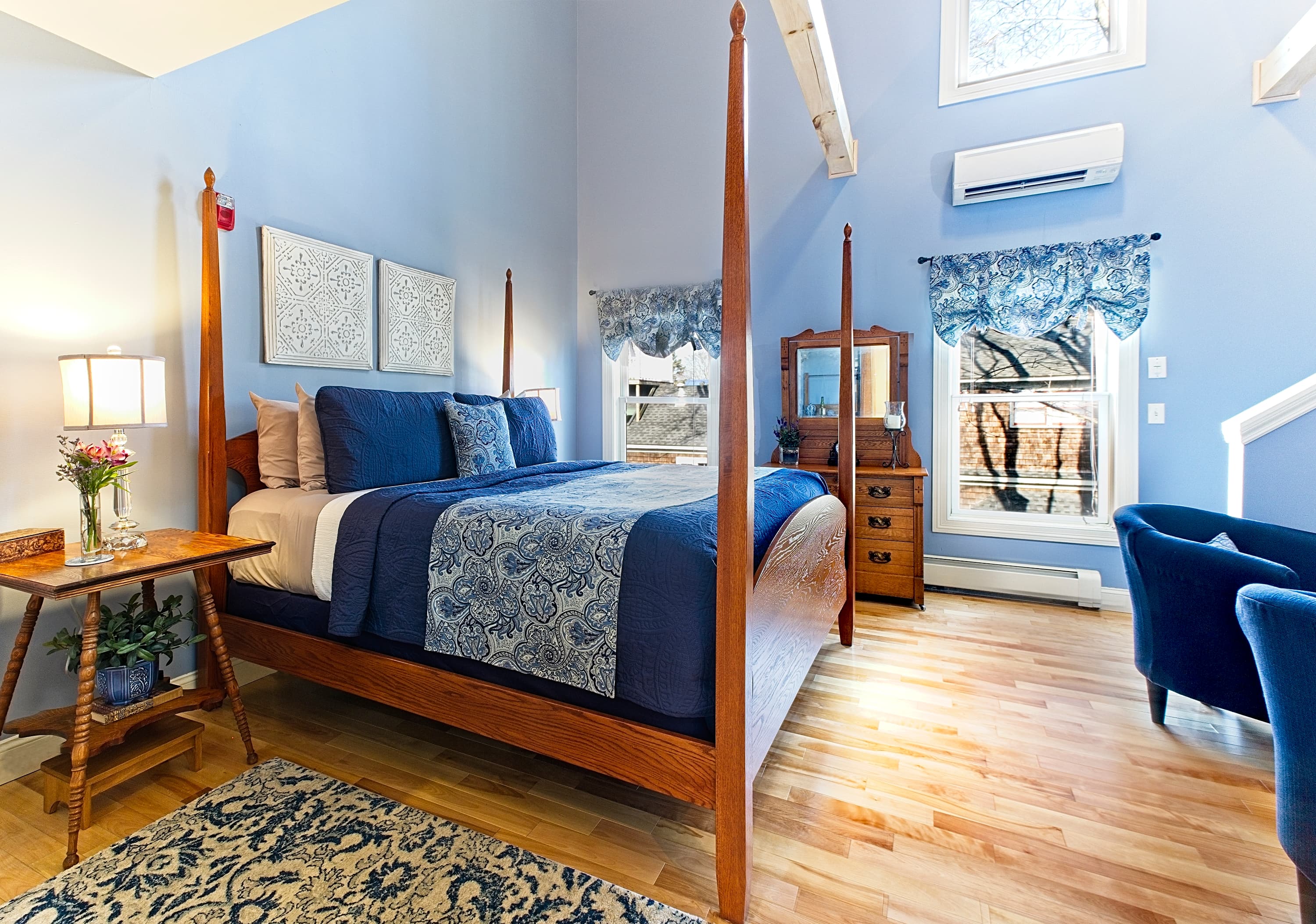 A sunlit luxury suite featuring a four-poster king bed with navy blue linens, light maple flooring, blue walls, and high ceilings with large windows and white structural beams.