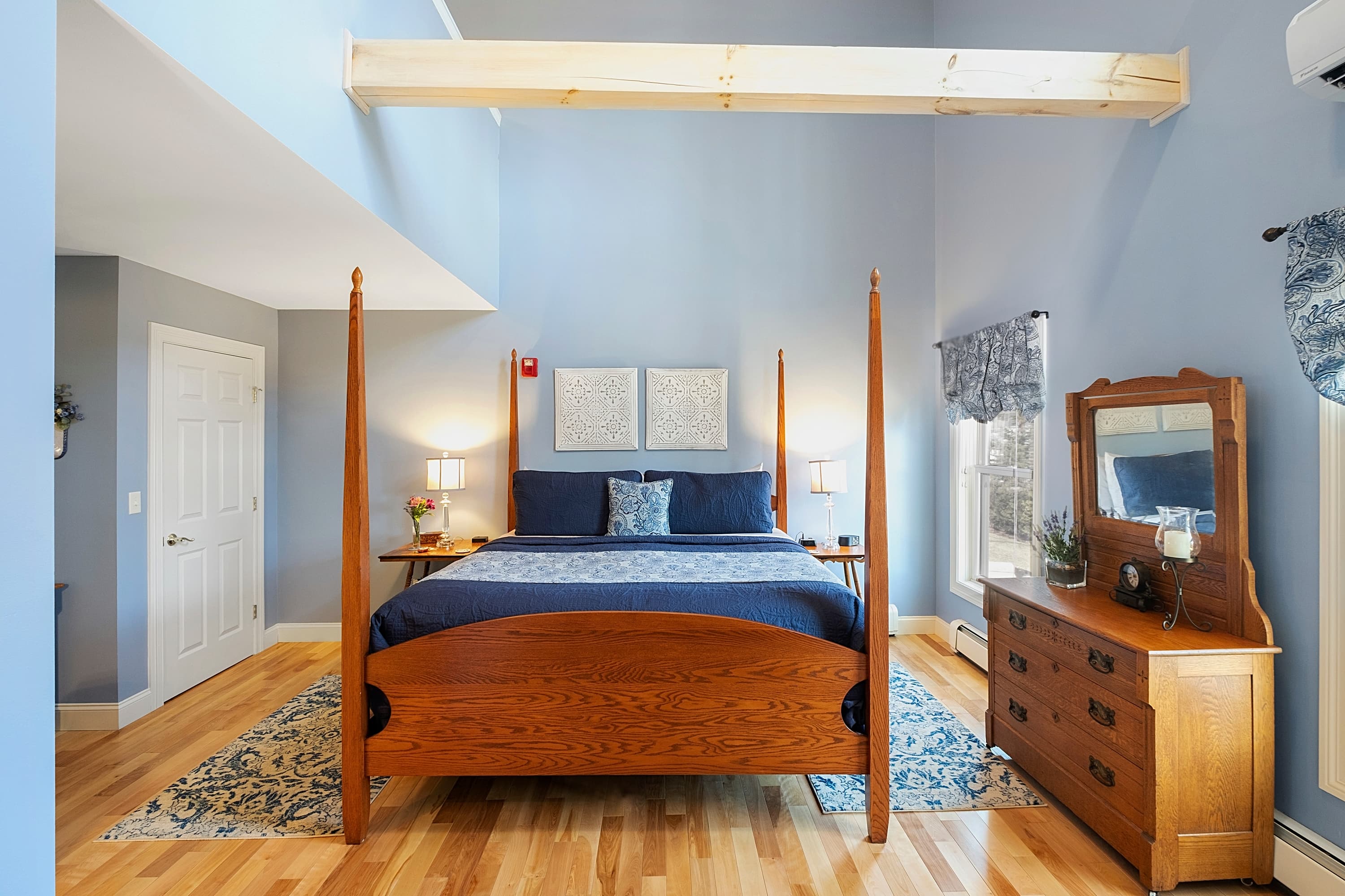Spacious luxury guestroom featuring a wooden four-poster king bed with navy blue linens, light hardwood floors, high ceilings with an exposed timber beam, and a matching wooden dresser.
