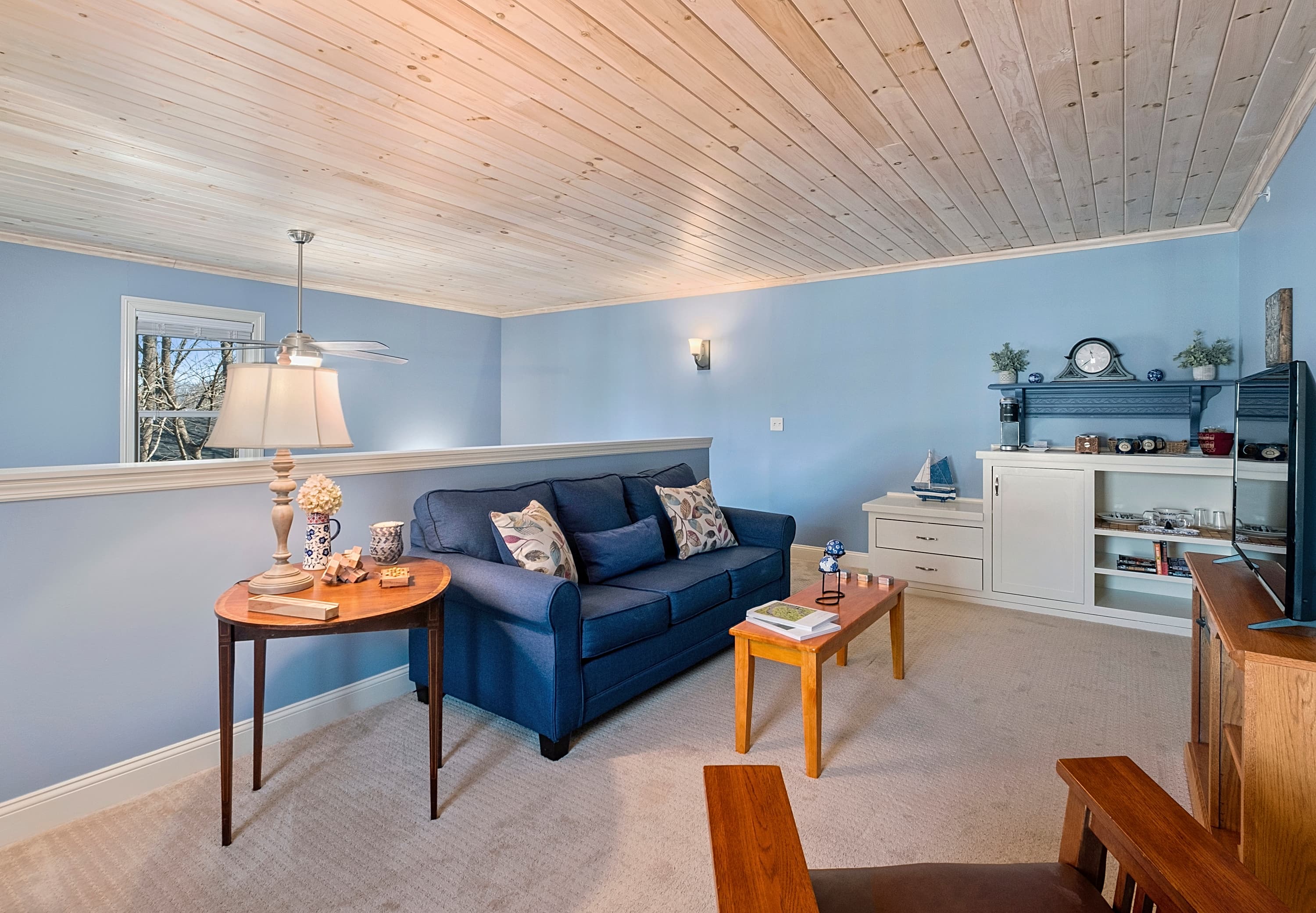 A secondary view of a loft sitting area featuring a blue sofa, a wooden coffee table, and a built-in refreshment center under a light-wood plank ceiling with light-blue walls.