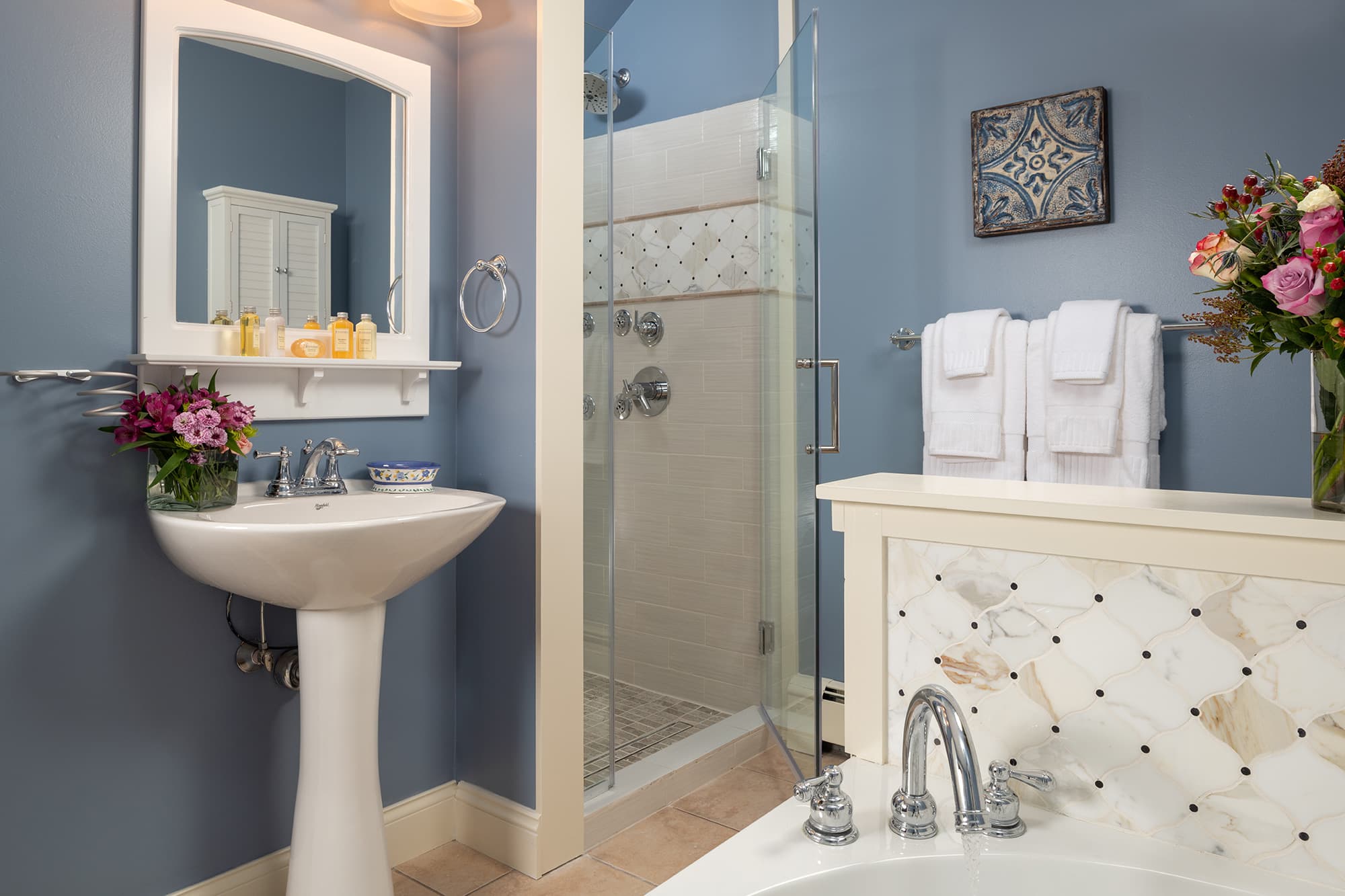 A luxurious en-suite bathroom featuring a white pedestal sink, a glass-enclosed spa shower with decorative tiling, and a large whirlpool tub against blue walls with white trim.