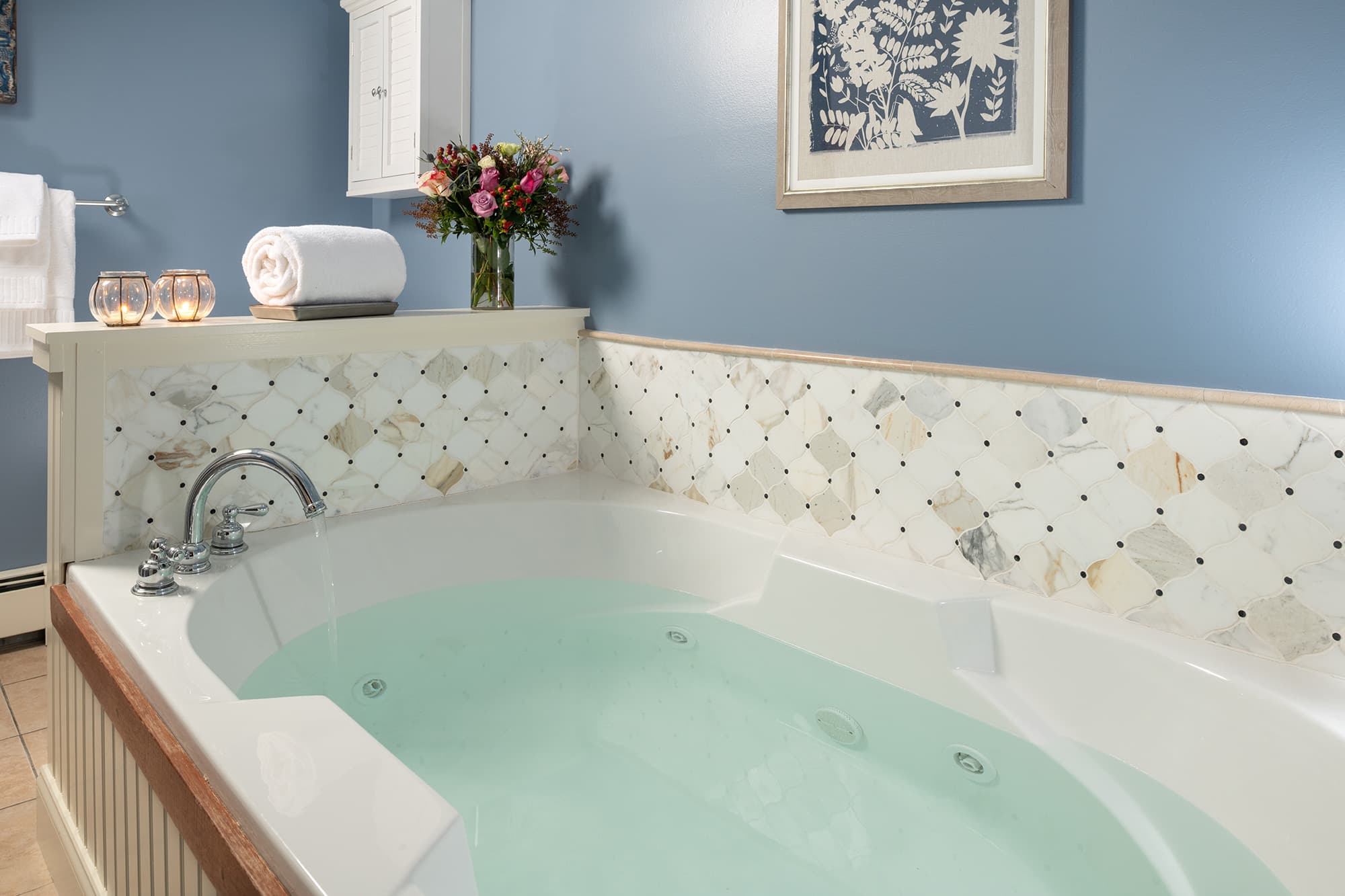 An oversized white whirlpool tub filled with water, featuring decorative diamond-patterned wall tiling, fresh flowers, and candles against a serene blue wall.