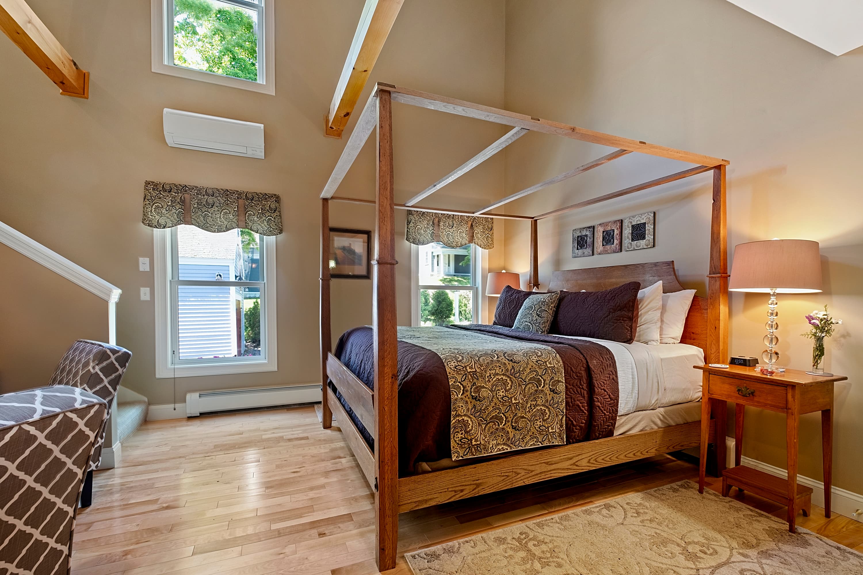 A sun-drenched luxury suite featuring a king-size wooden canopy bed with plum and patterned linens, light maple floors, high ceilings with exposed beams, and a glimpse of the staircase.