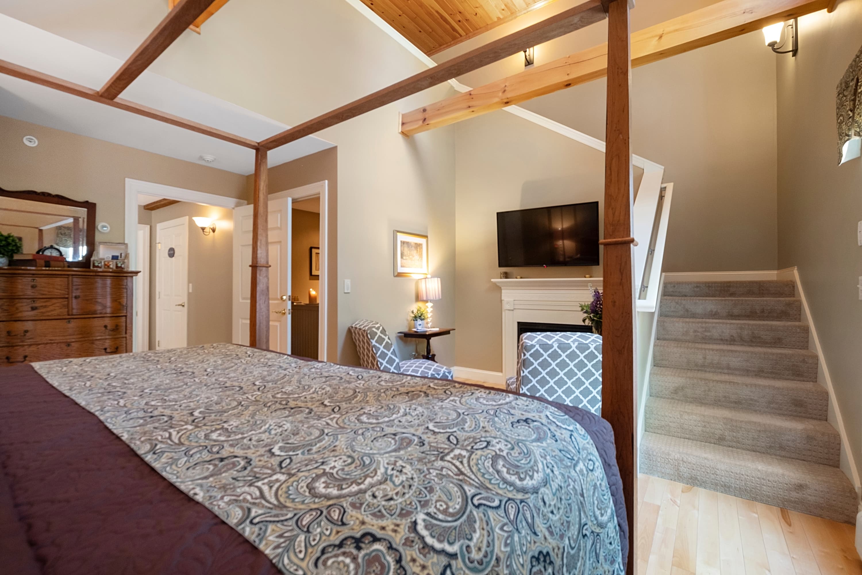 A spacious bedroom view showing a wooden canopy bed with paisley linens, a gas fireplace with a mounted TV, and a carpeted staircase leading to a loft level under high ceilings with exposed wood beams.