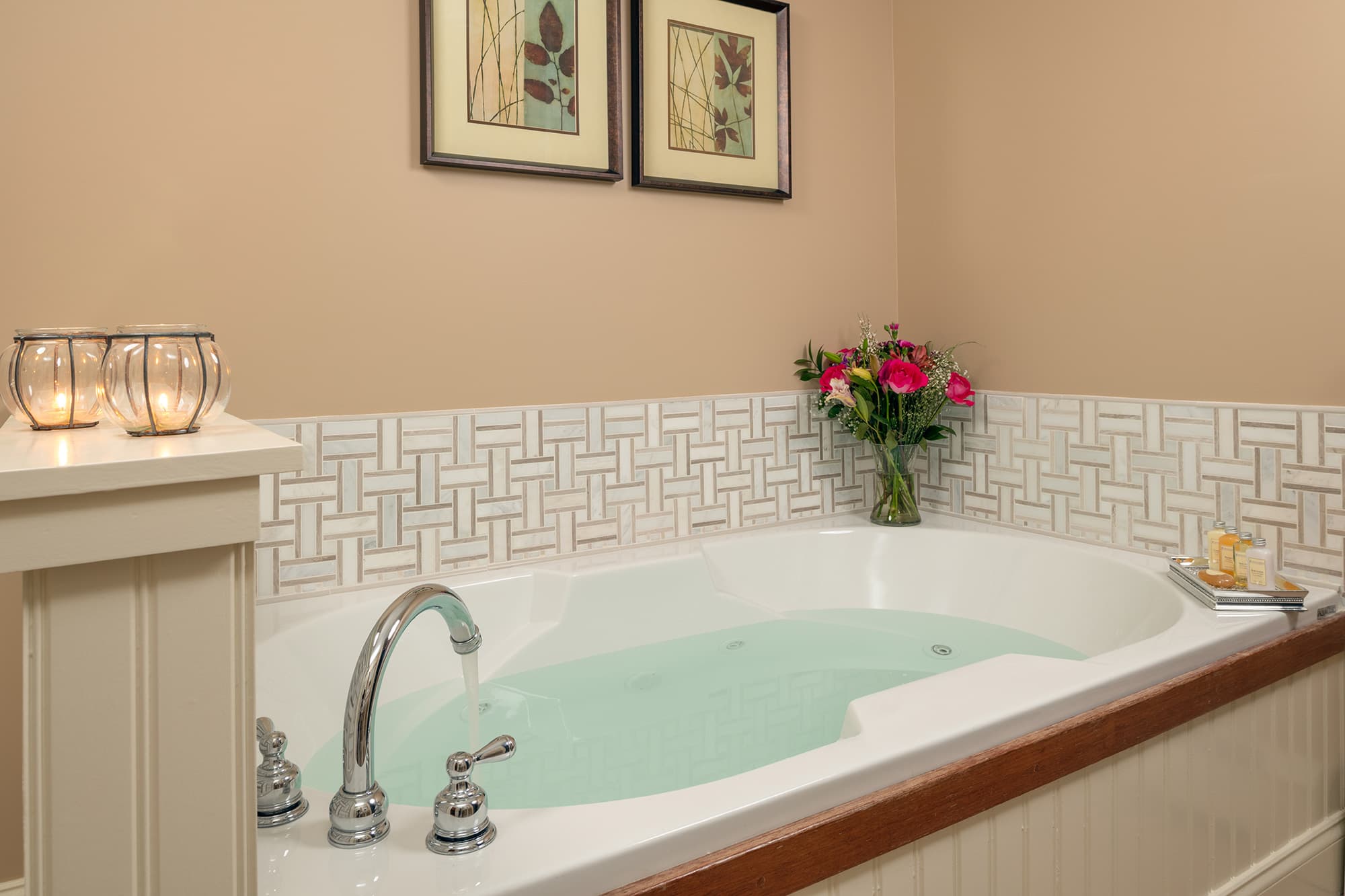 An oversized white whirlpool tub filled with water, accented by a basket-weave tile backsplash, fresh pink flowers, and glowing candles against warm tan walls.