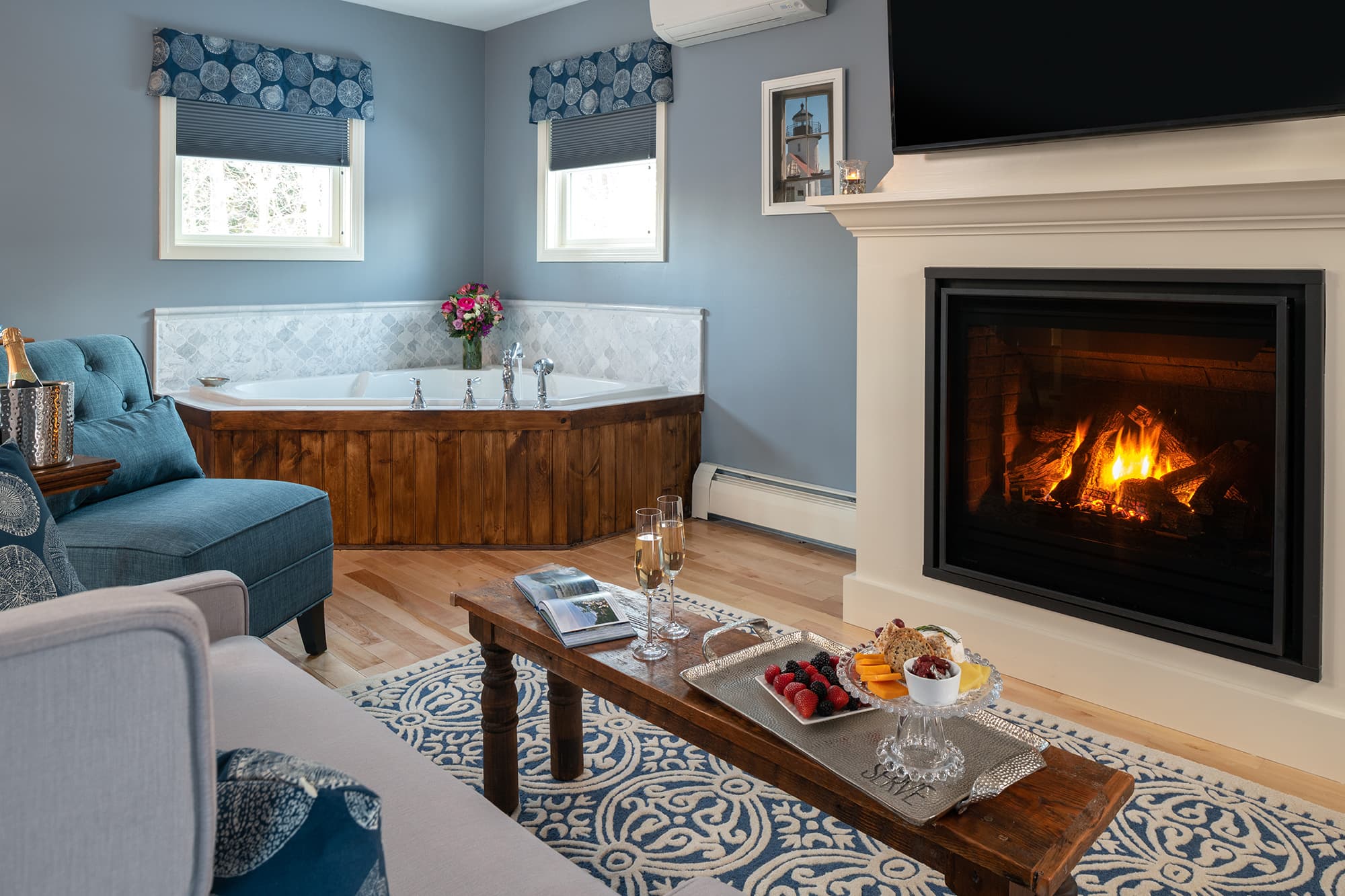 A cozy sitting area in a luxury suite featuring a warm gas fireplace, a wooden coffee table with a snack tray, and a large triangular whirlpool tub tucked into a sunlit corner with slate blue walls.