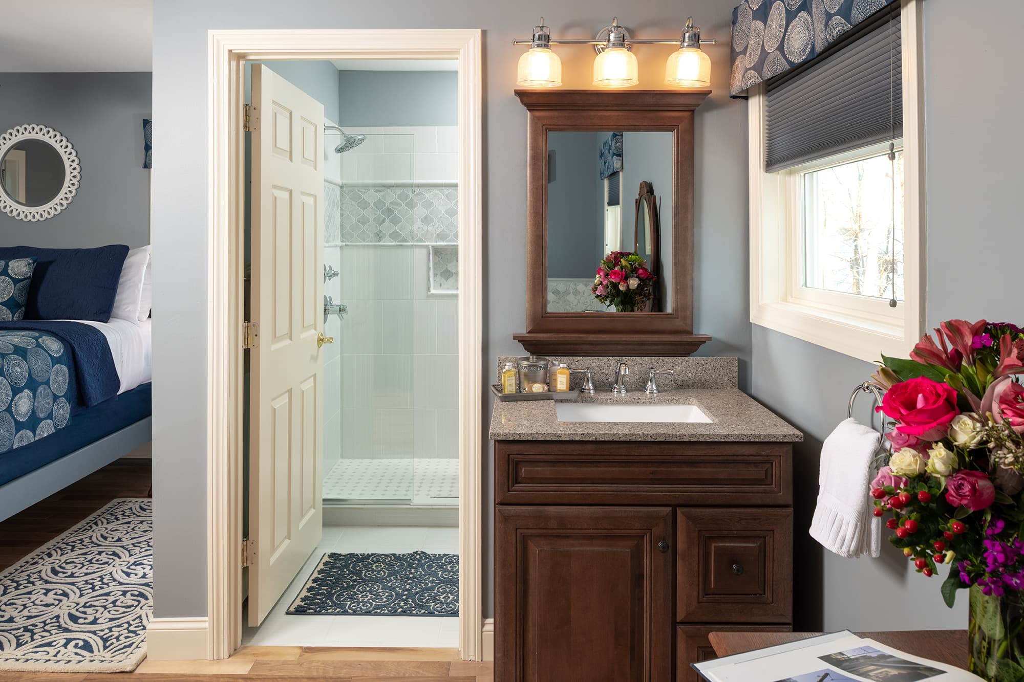 A sophisticated en-suite bathroom area featuring a dark wood vanity with a stone countertop, a matching framed mirror, and a view into a bright glass-enclosed spa shower with mosaic tile accents.