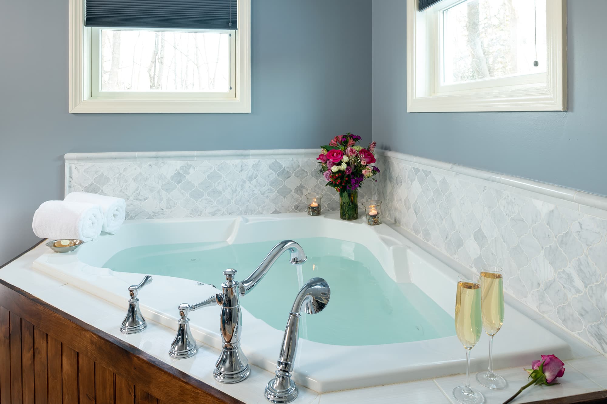 A close-up of an oversized triangular whirlpool tub filled with water, featuring a white marble tile backsplash, two champagne flutes, a bouquet of flowers, and candles in a sunlit corner.