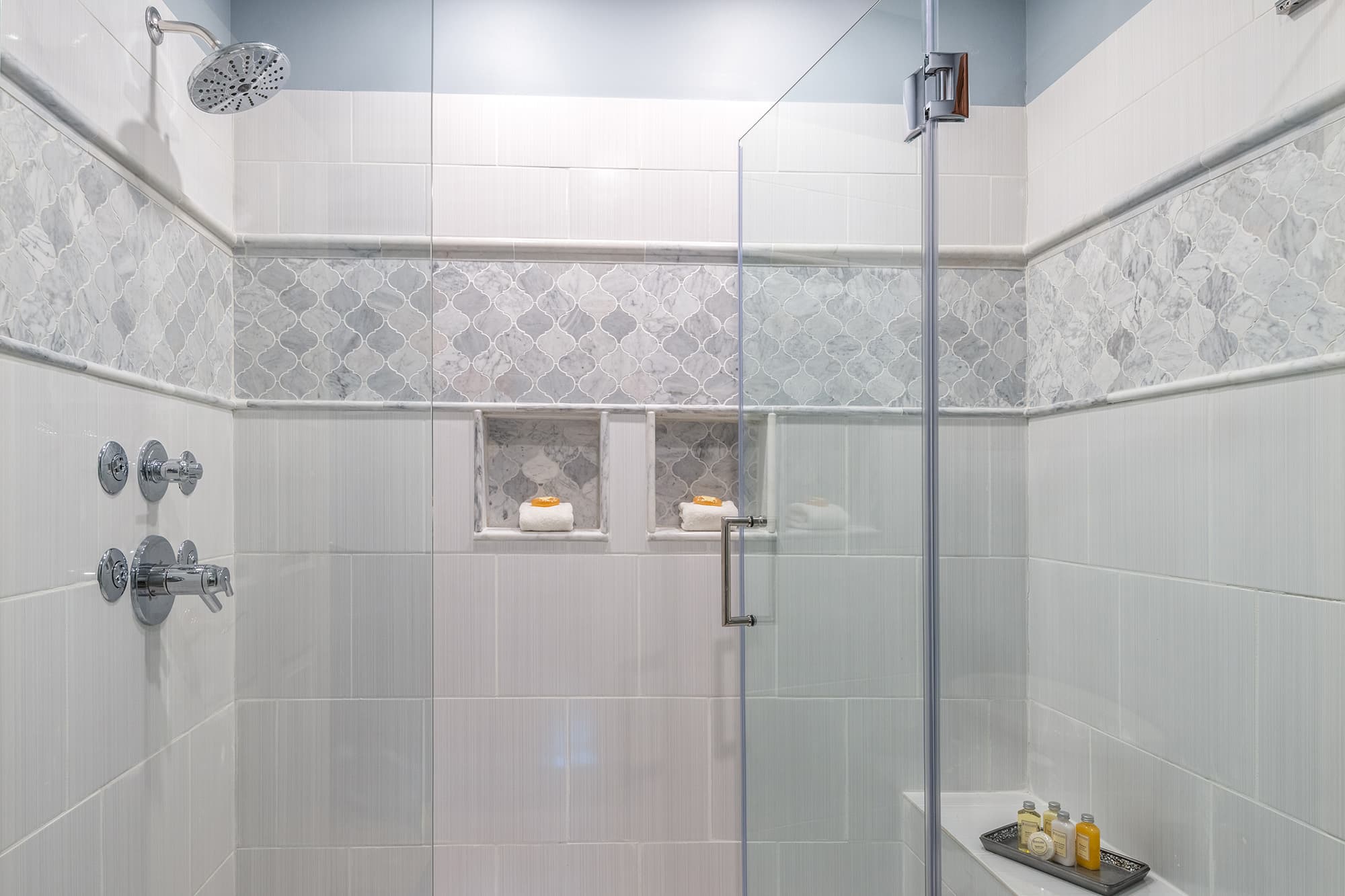 A large glass-enclosed spa shower featuring white subway tiles, a decorative mosaic tile border, dual built-in shelving nooks, and luxury massage body sprays.