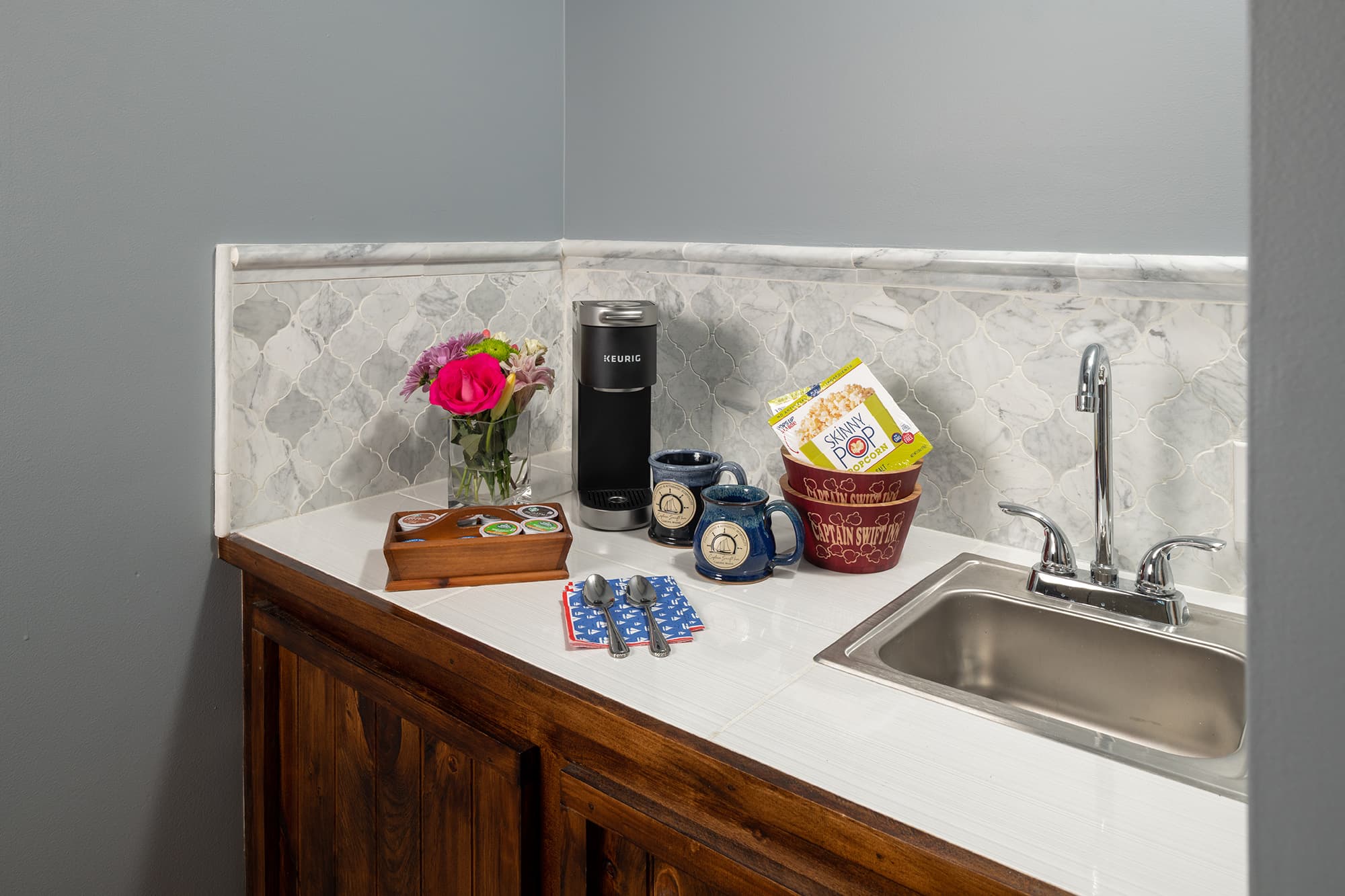 A private wet bar area featuring a white countertop with a small stainless steel sink, a Keurig coffee maker, branded mugs, and snacks, set against a marble tile backsplash and slate blue walls.