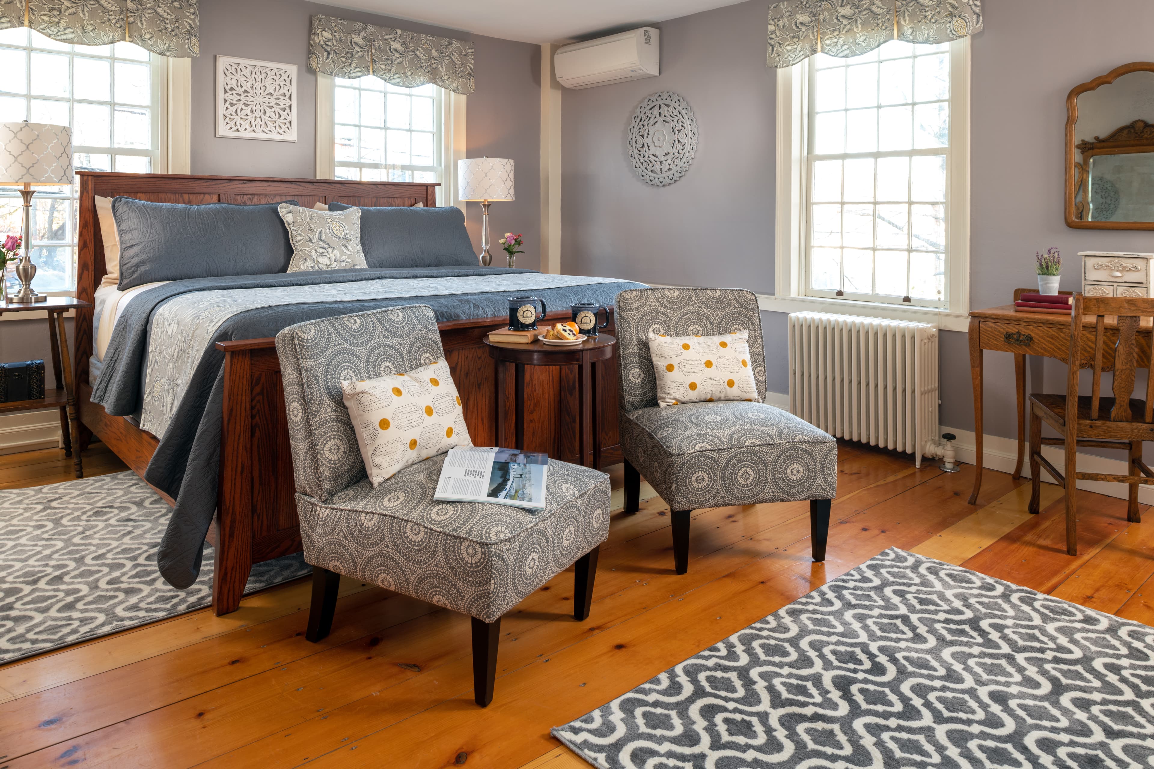 A classic, sun-filled guestroom featuring a king-size sleigh bed, wide-plank pumpkin pine flooring, two patterned slipper chairs with a small side table, and three large original windows with decorative valances.
