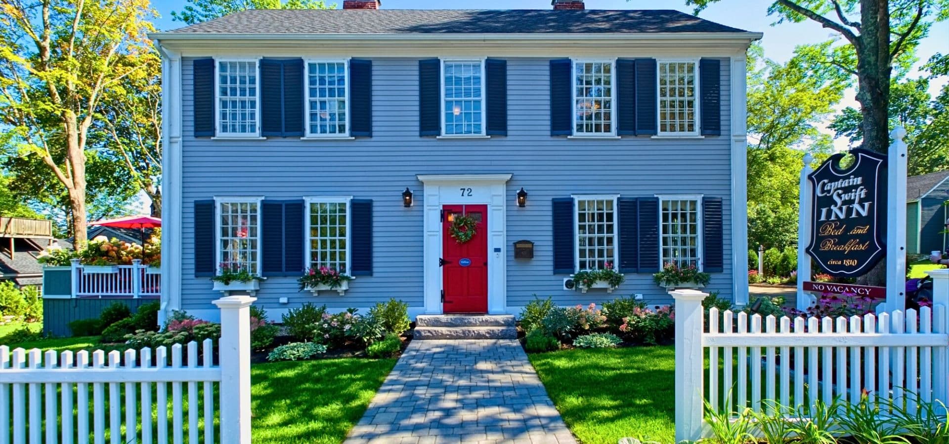 A charming blue inn with white shutters and a red front door, surrounded by lush greenery and a white picket fence.