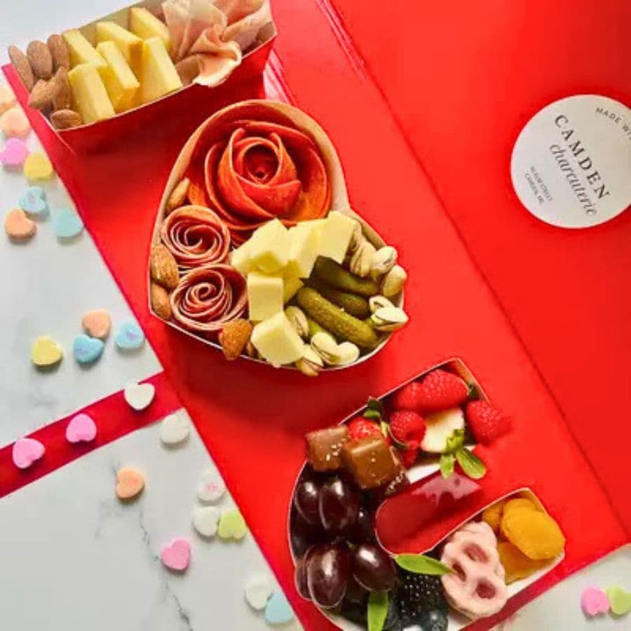 A colorful assortment of cheeses, meats, fruits, and treats arranged in heart-shaped containers on a red background.