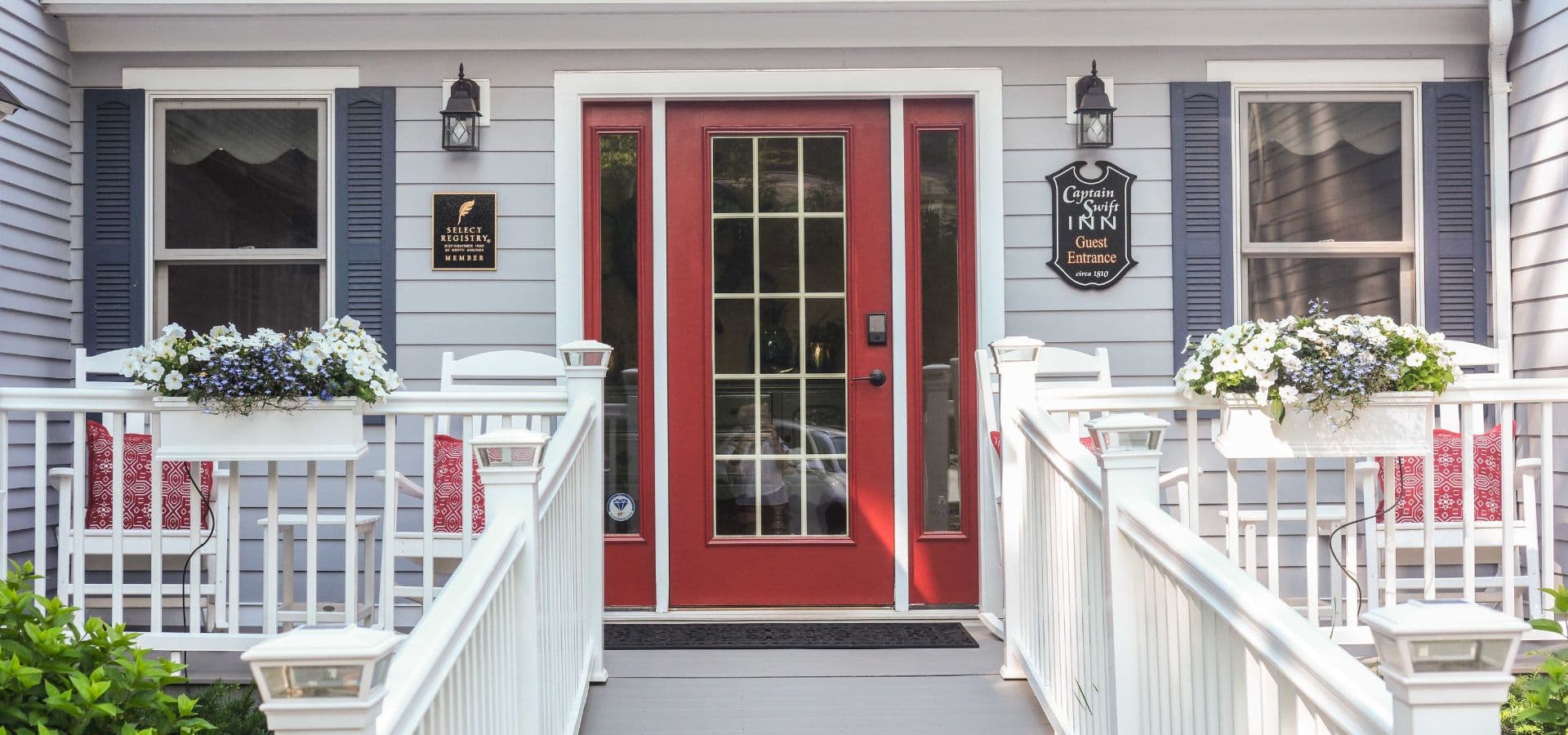 Front entrance of a light gray inn with a red door, flanked by flower boxes and a welcoming sign.
