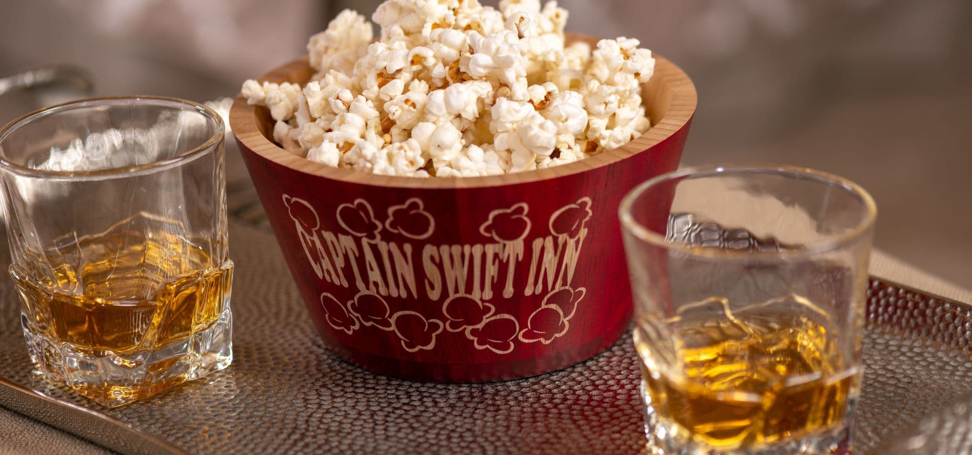 A bowl of popcorn sits between two glasses of whiskey on a decorative tray.