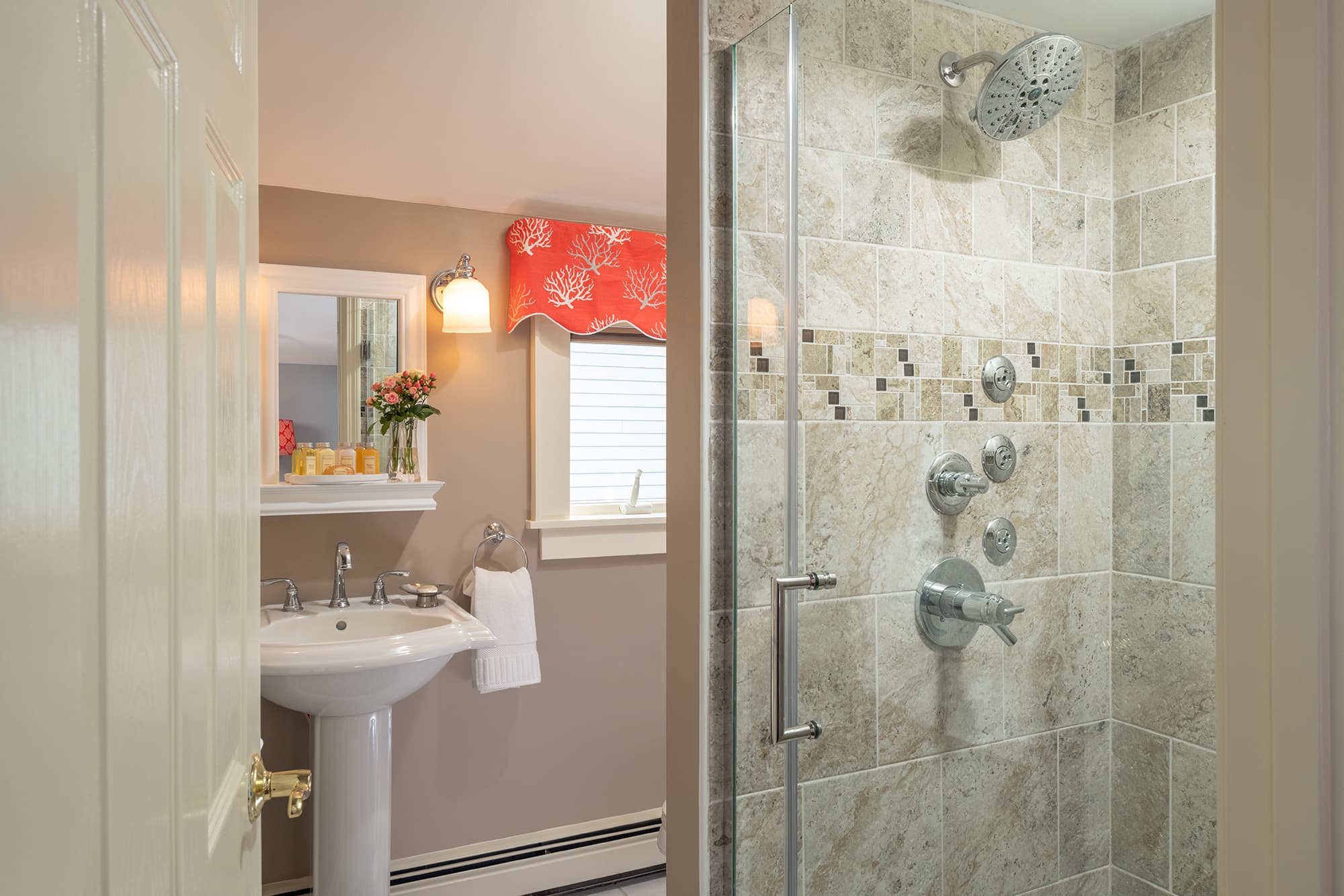 A bright en-suite bathroom featuring a glass-enclosed walk-in shower with natural stone tiles and luxury body sprays, next to a white pedestal sink and a window with a vibrant coral valance.