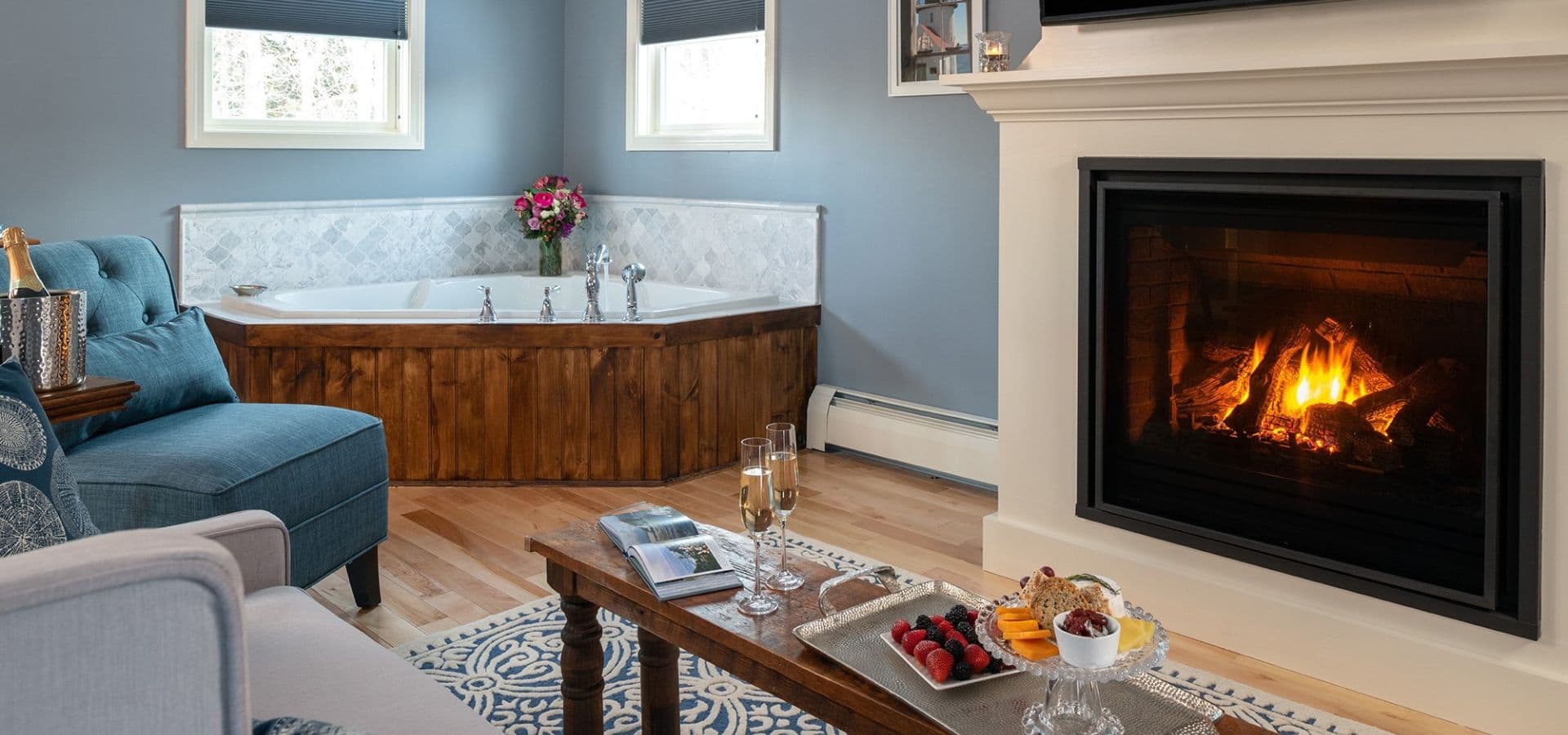 A cozy living space featuring a fireplace, a blue loveseat, a wooden bathtub, and a coffee table with snacks and drinks.