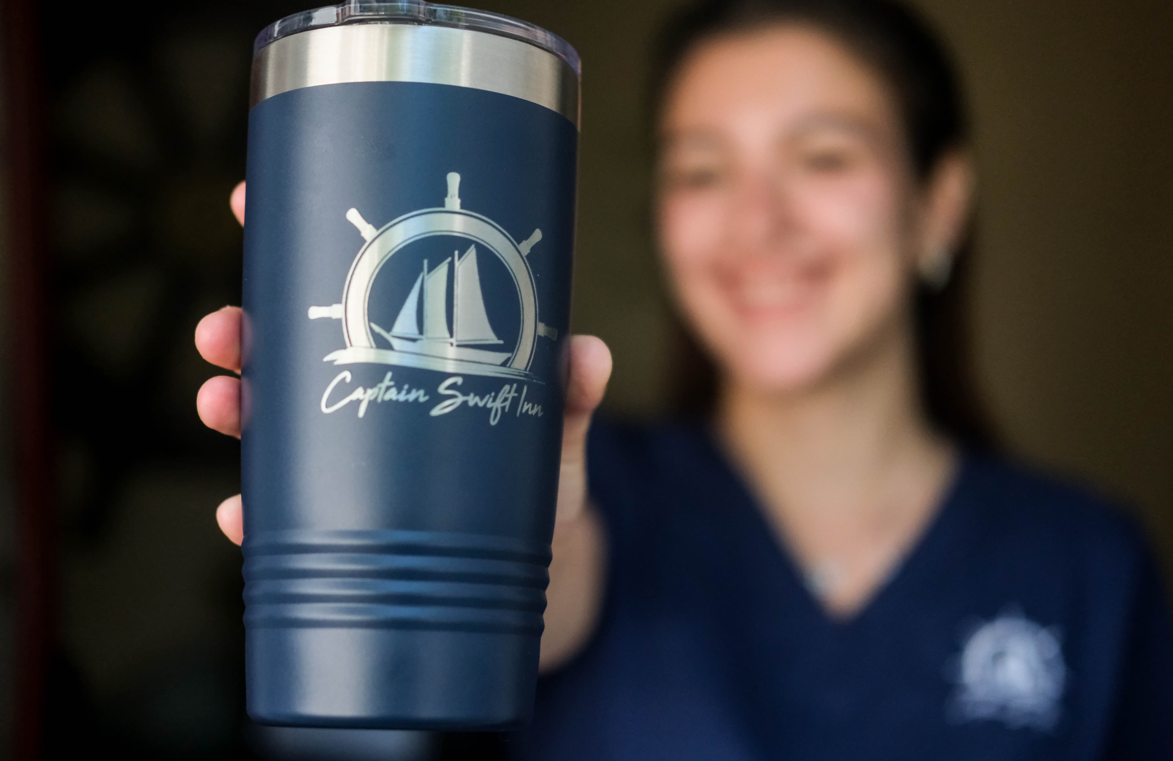 A person holds a navy blue tumbler featuring a sailing design and the text "Captain Swift Inc." while smiling in the background.