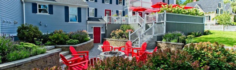 A cozy outdoor seating area with red chairs and umbrellas surrounded by lush gardens and a blue house.