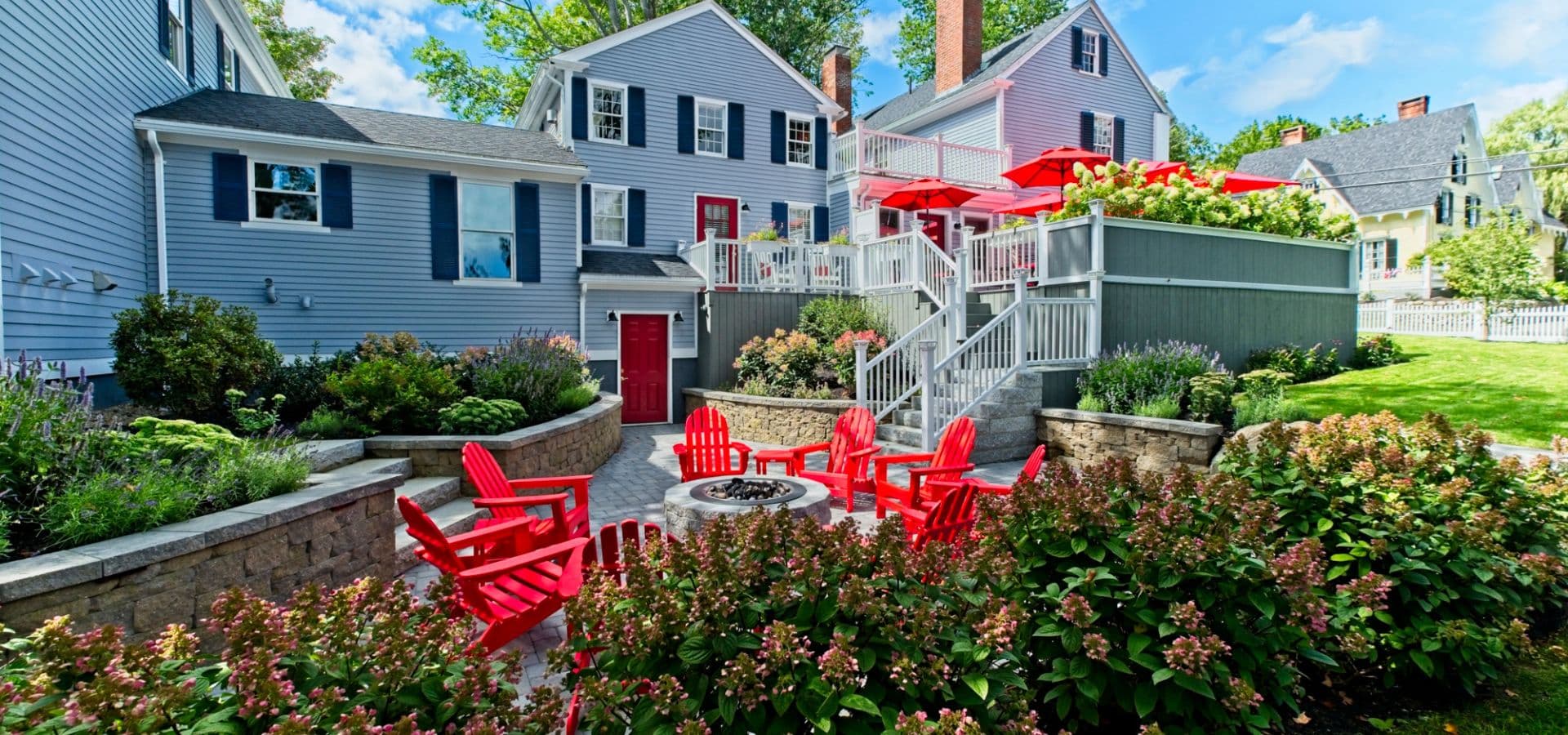 A landscaped backyard features red Adirondack chairs surrounding a fire pit, with a deck and blooming flowers in the background.