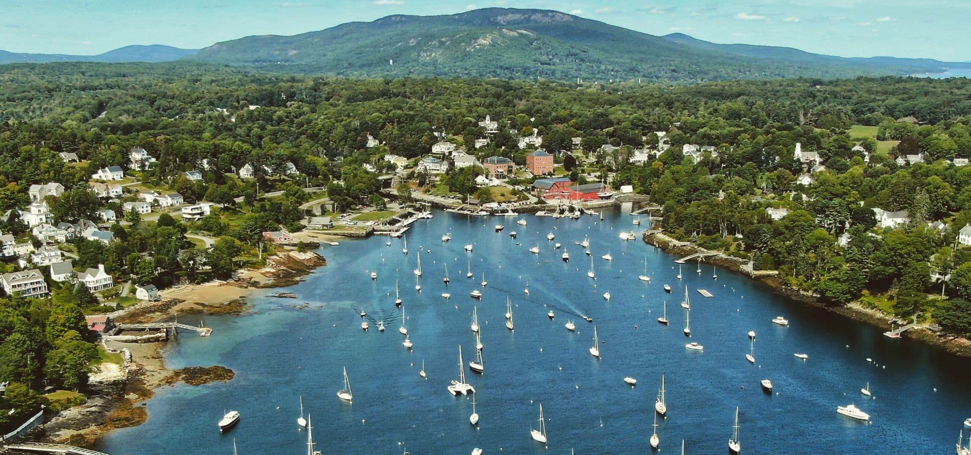 Aerial view of a scenic harbor surrounded by green hills and dotted with sailboats.