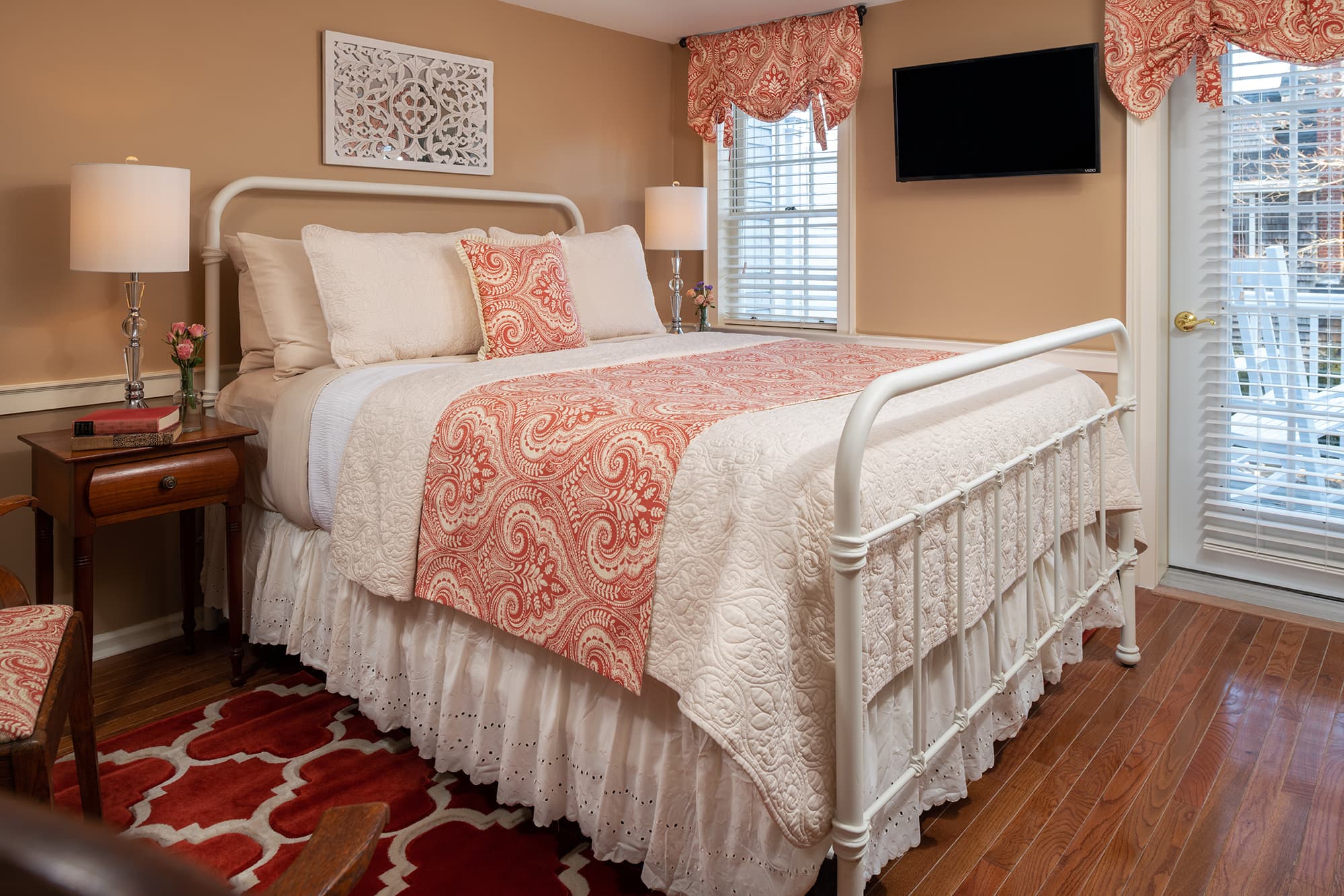 A charming guestroom featuring a white iron bed with coral and cream paisley linens, warm tan walls, and hardwood floors. The room includes a mounted flat-screen TV and a glass door leading to a private outdoor deck.