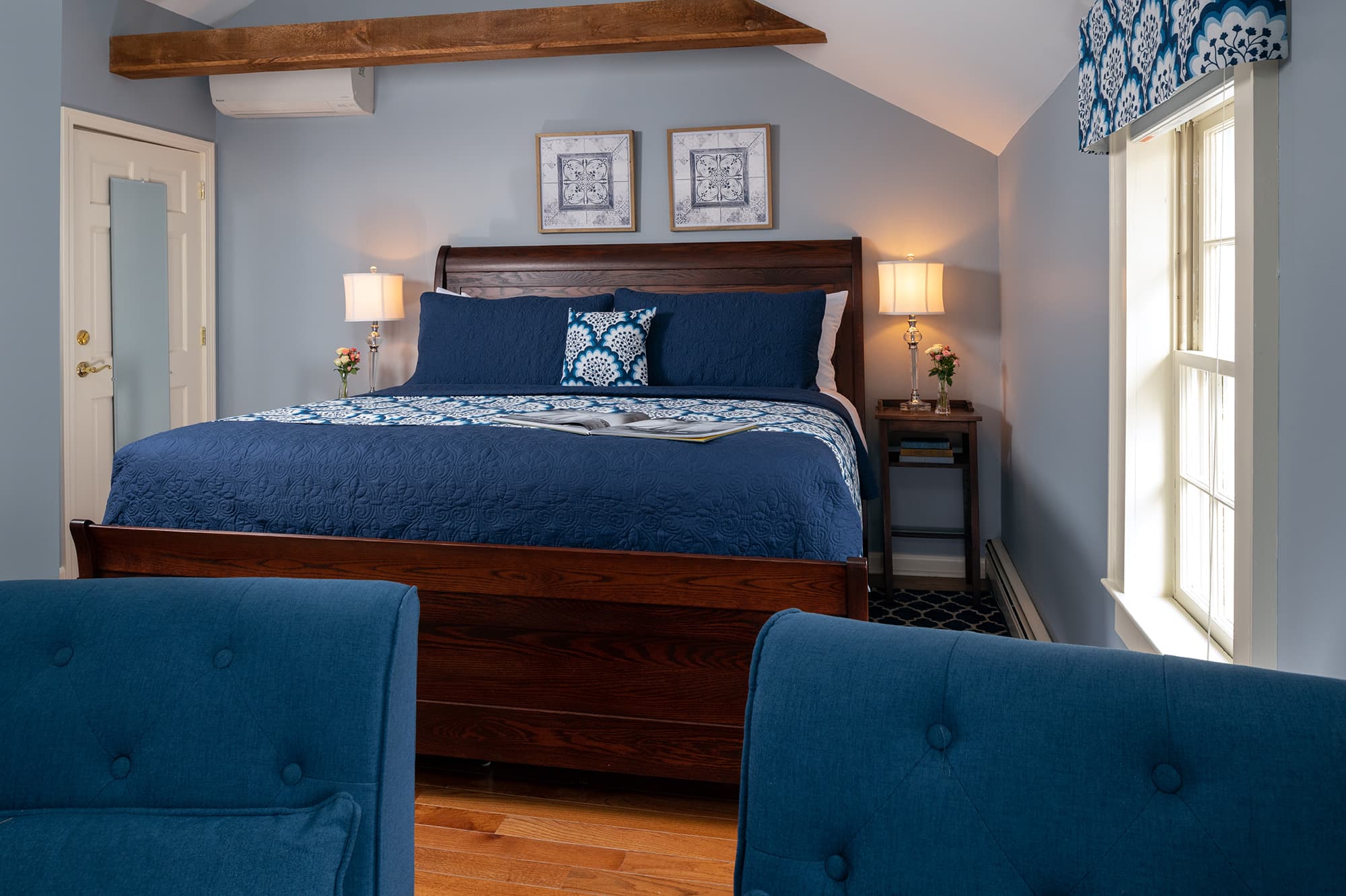 A serene guestroom featuring a king-size wooden sleigh bed with navy blue linens, set under a vaulted ceiling with a prominent exposed wood beam and light blue walls.