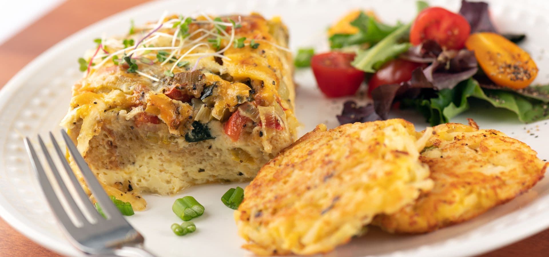 A plate of baked egg casserole with two golden hash browns and a side salad featuring cherry tomatoes and mixed greens.