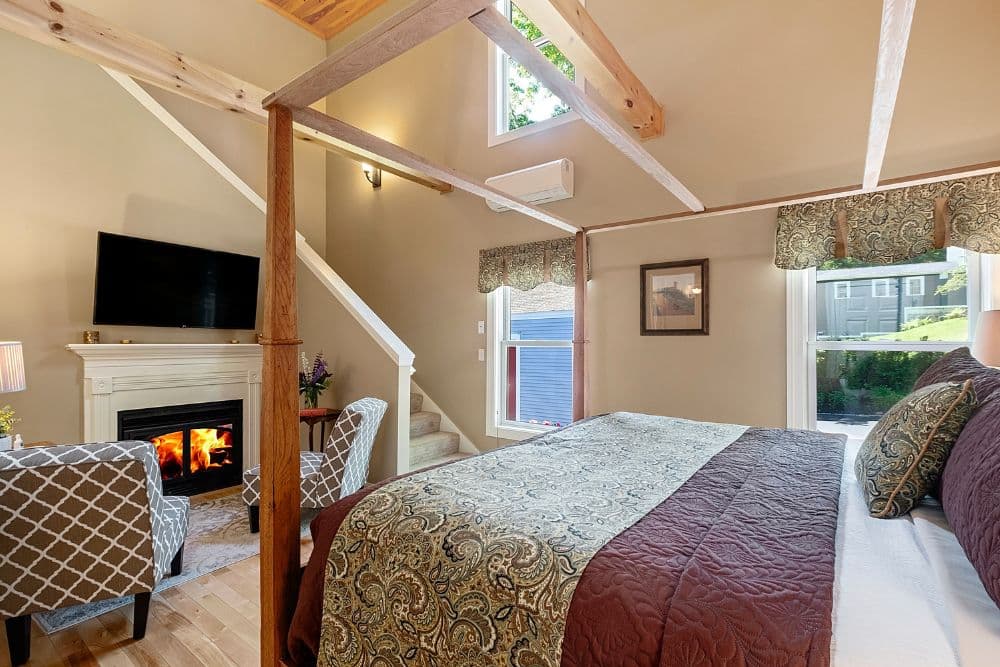 A cozy bedroom featuring a decorative bed, fireplace, comfortable seating, and large windows that allow natural light.
