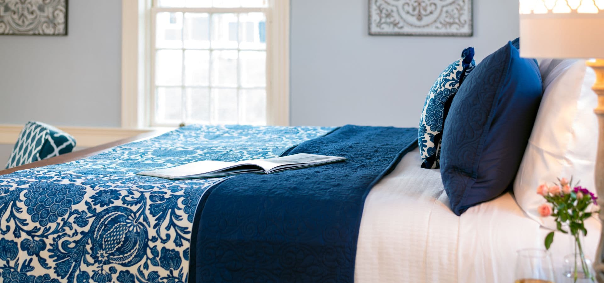 Stylishly arranged bed with blue and white bedding, a book, and decorative pillows in a serene room.