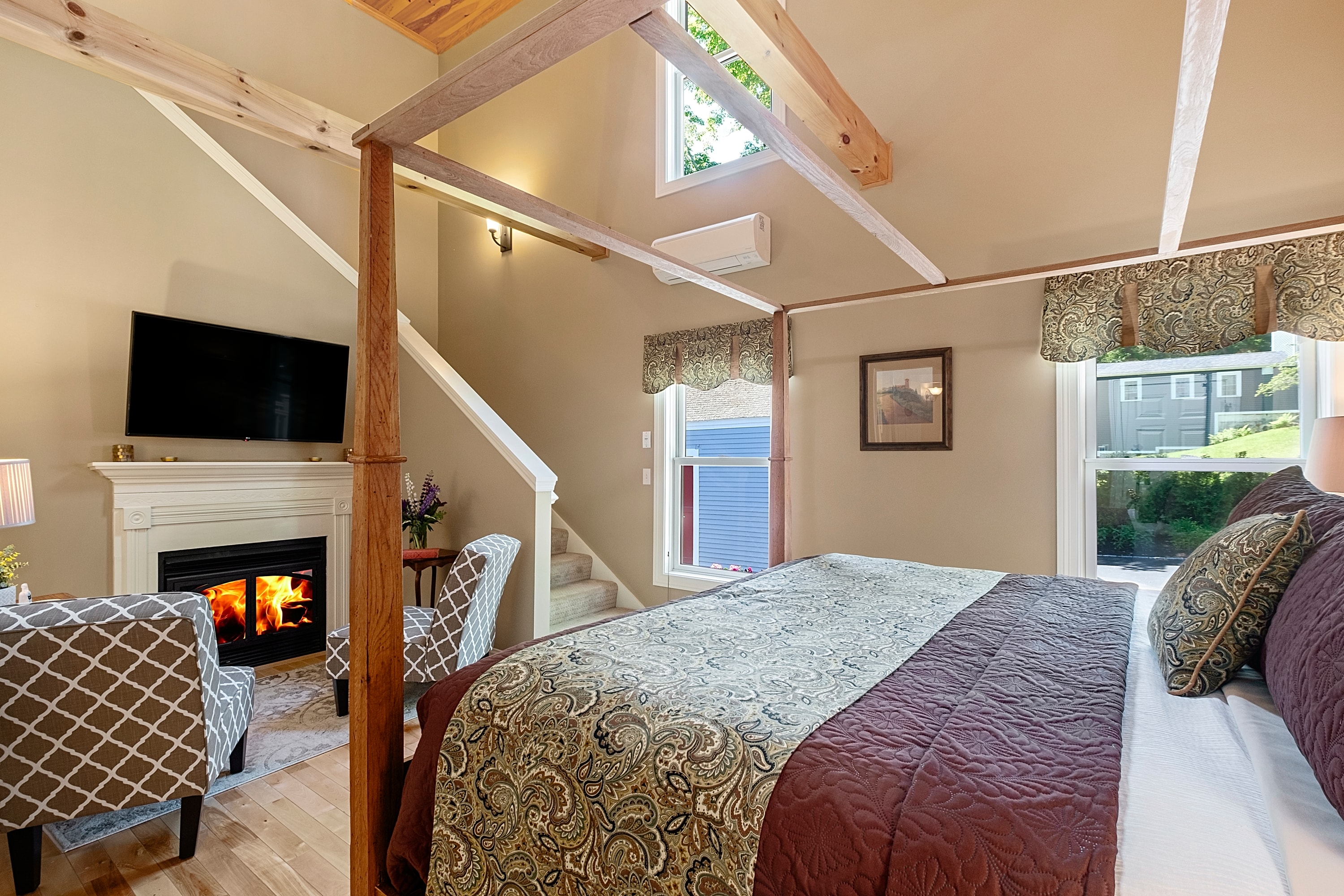 A warm, two-level luxury suite featuring a wooden canopy bed with patterned linens, a gas fireplace with a flat-screen TV above, tan walls, and a staircase leading to a sunlit loft.
