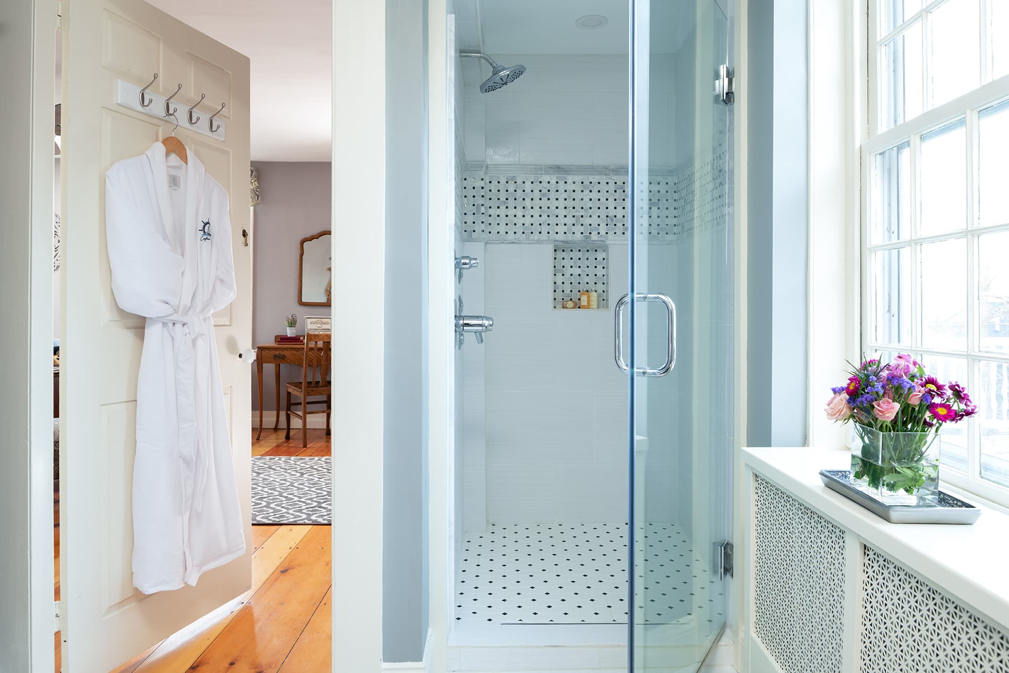 A bright en-suite bathroom featuring a glass-enclosed walk-in shower with mosaic tile accents, a white plush robe hanging on the door, and a large window providing natural light.