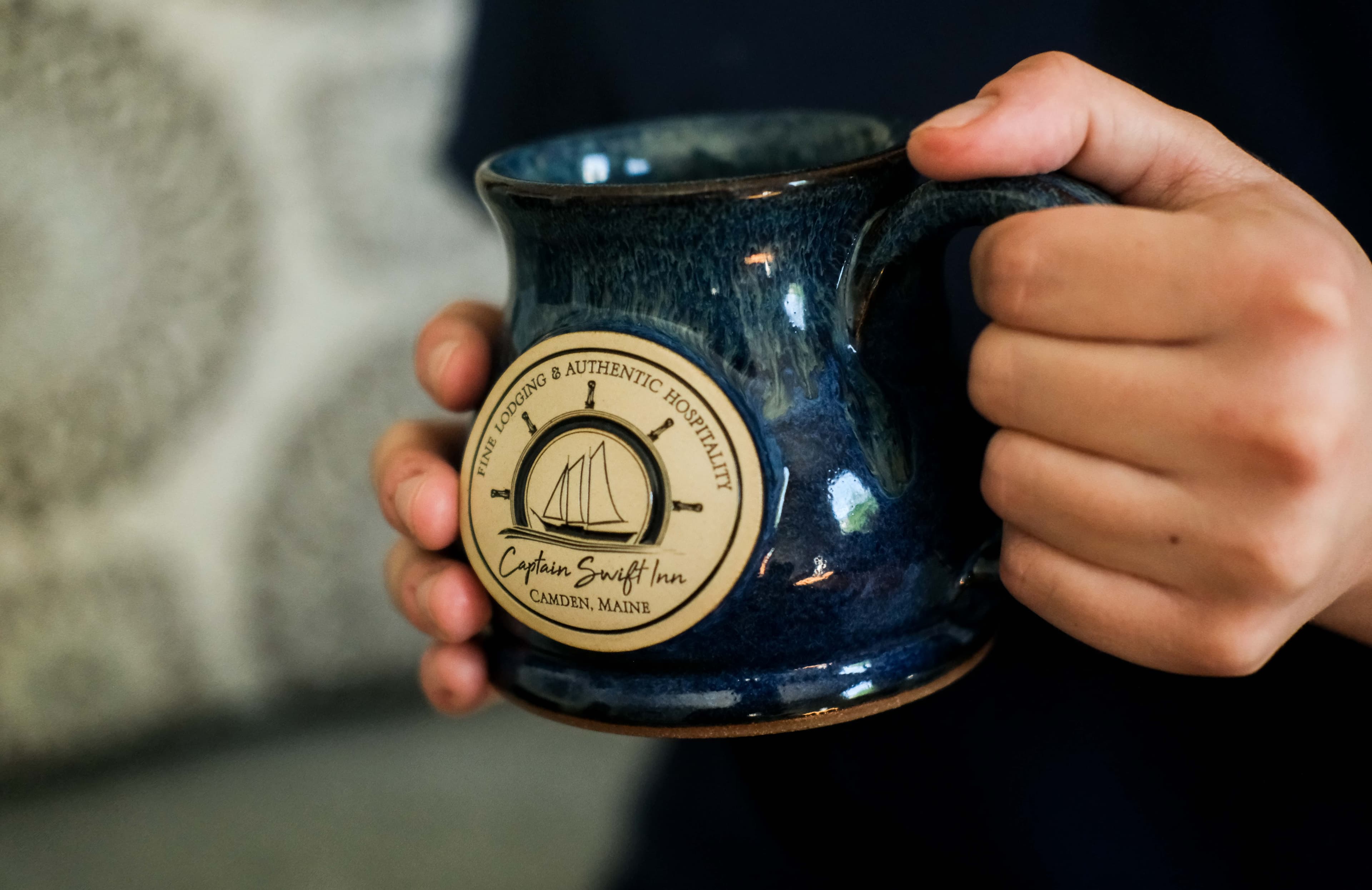A person holds a blue mug with a logo for Captain Swift Inn, Camden, Maine.
