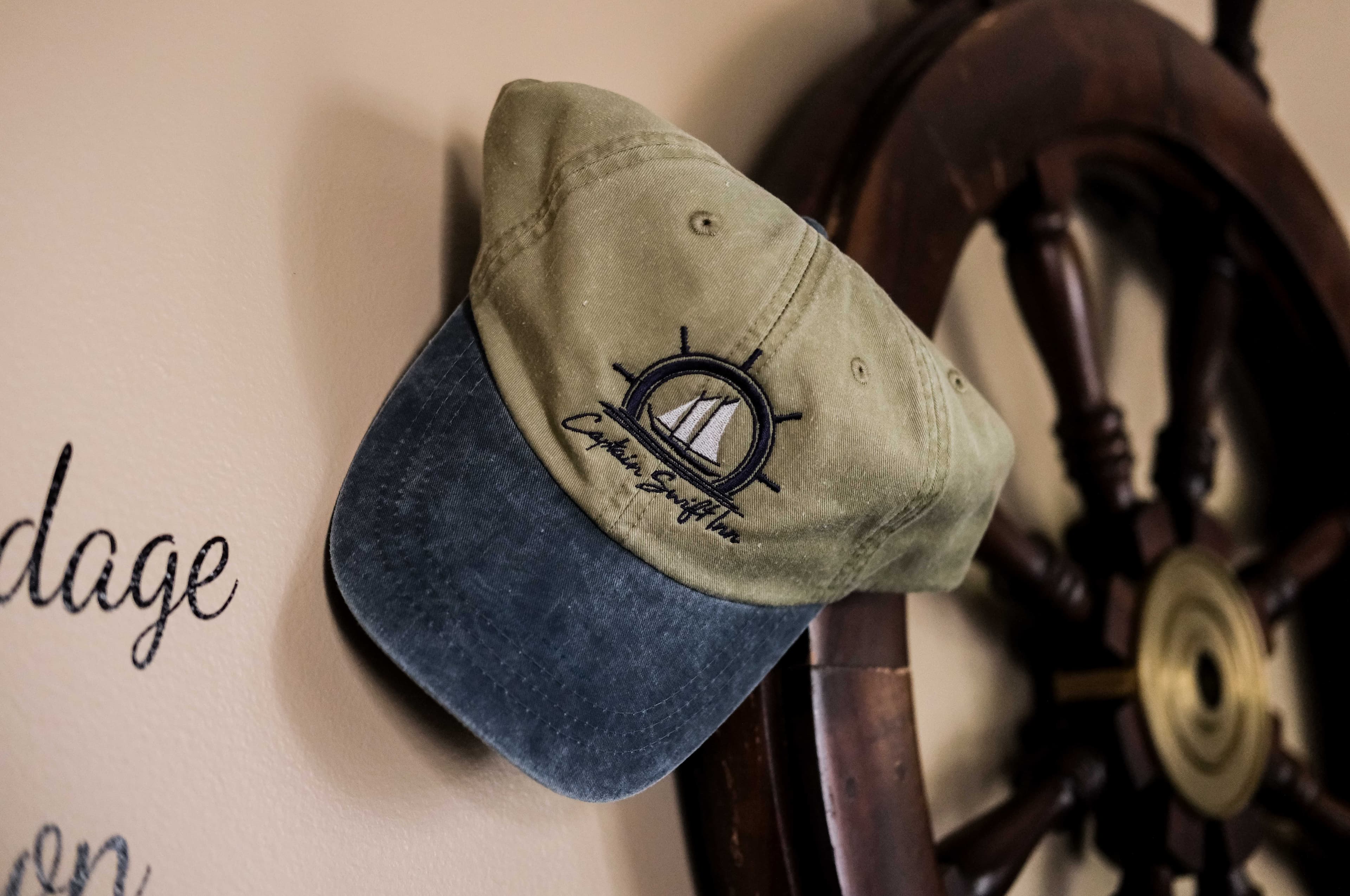 A green and blue cap with a nautical design is hung on a wall next to a wooden ship's wheel.