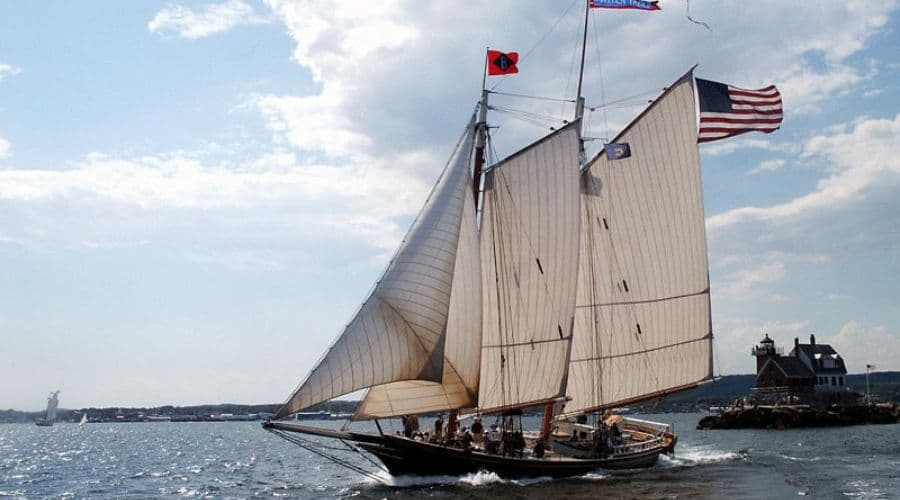 A sailing ship with multiple sails navigates calm waters under a partly cloudy sky.