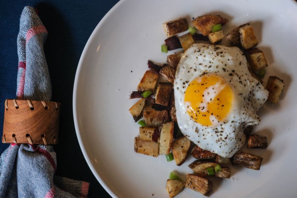 A fried egg sits atop seasoned roasted potatoes, garnished with green onions, on a round white plate.