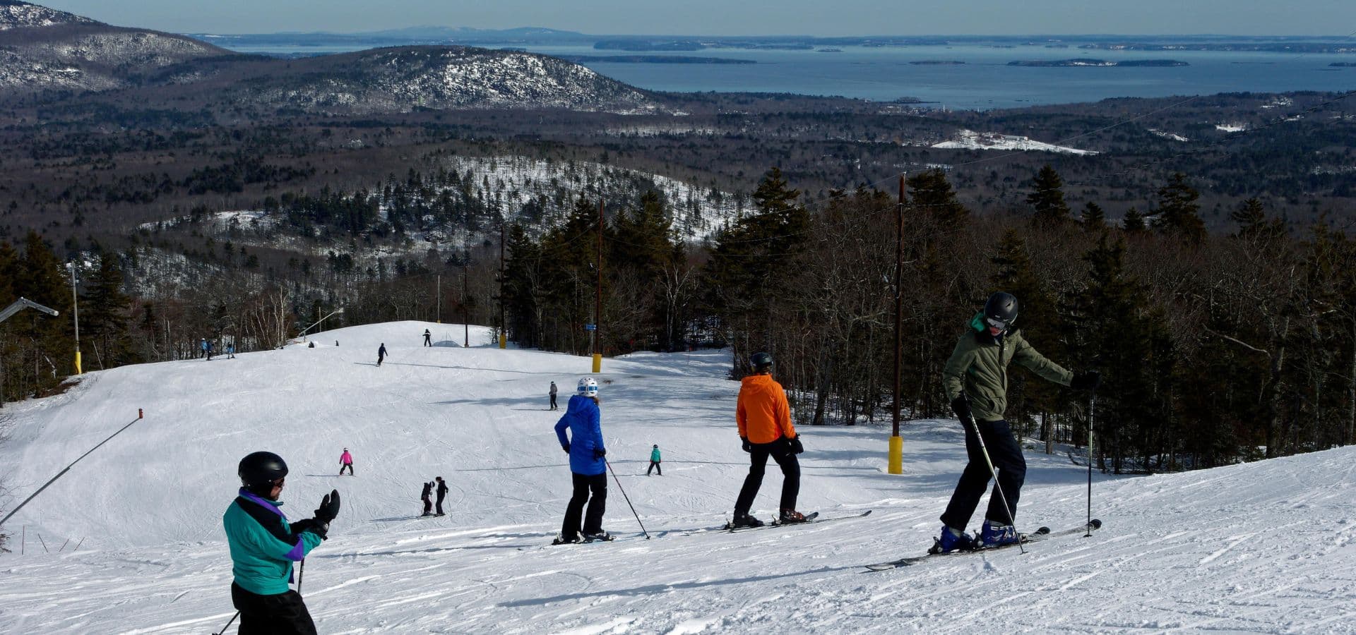 Skiers navigate a snowy slope with a scenic view of mountains and a lake in the background.