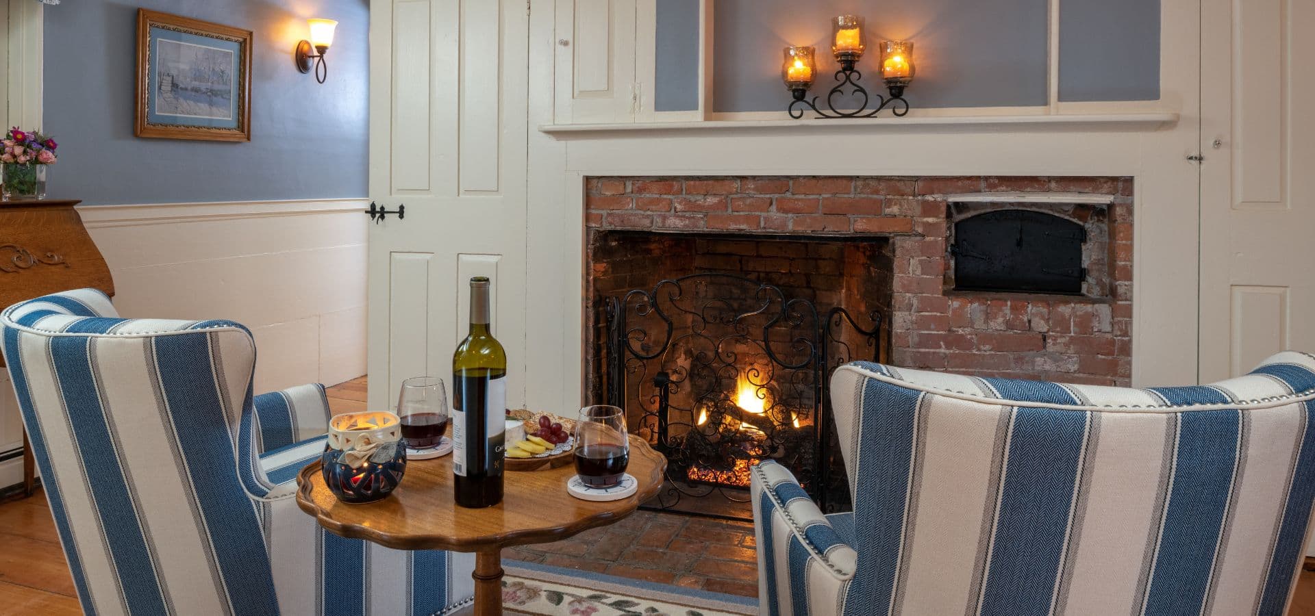 A cozy living room featuring two striped chairs, a wooden table with wine, snacks, and a warm fireplace.
