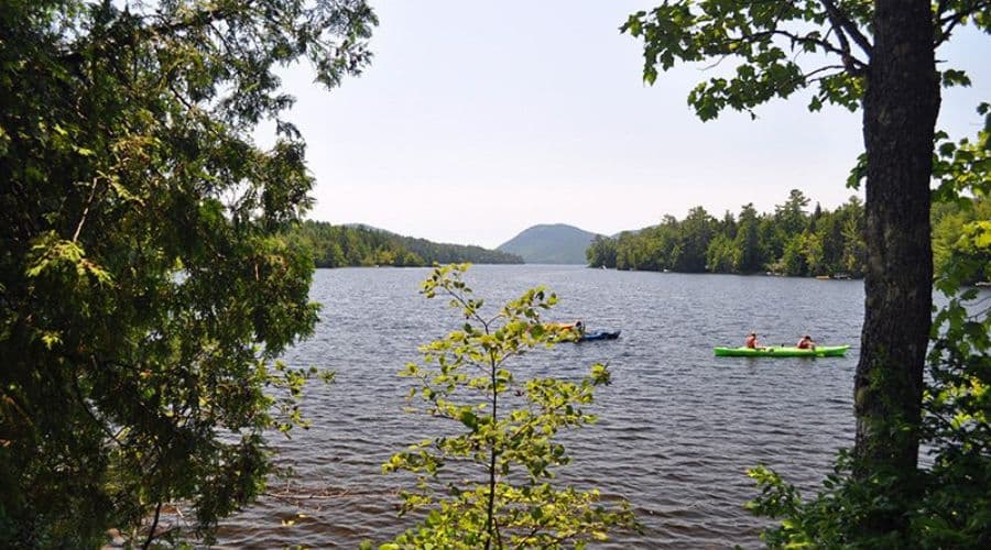 Two kayakers paddle on a calm lake surrounded by trees and mountains.